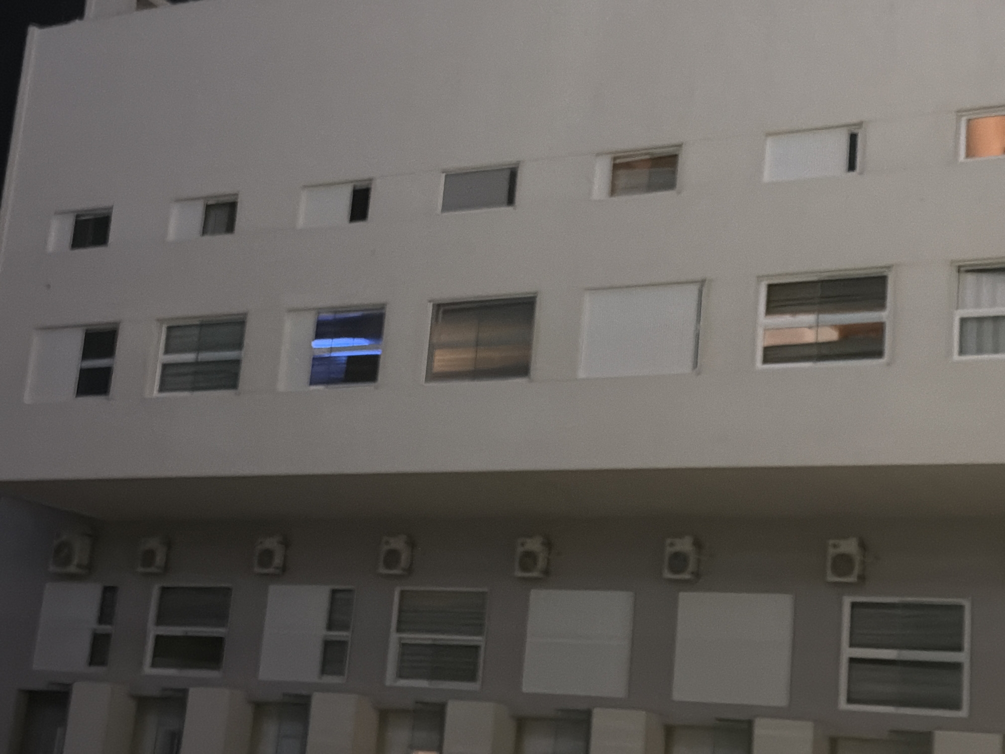 The image shows a nighttime exterior view of a multi-story apartment building, likely in Budva, Montenegro. The building has a light-colored facade and is divided into sections. On the left side of the image, there are balconies with railings and visible air conditioning units mounted on the walls between windows. The windows themselves vary, with some featuring rolled-down shutters, some with curtains, and others appearing to be dark. On the right side of the building, the facade is smoother, with more windows visible. Some of these windows also have shutters, while others show faint interior lights or reflections. The overall impression is a residential building at night, with minimal activity visible.