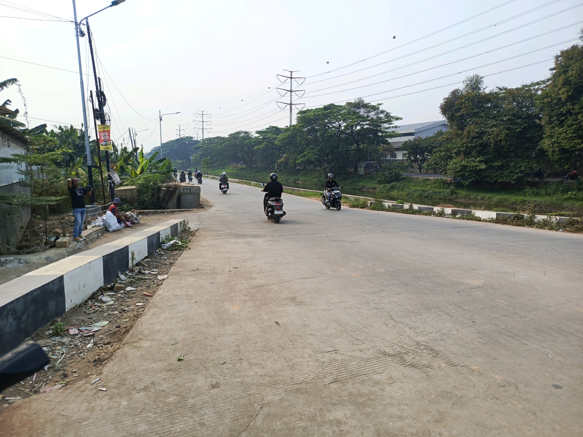 The image depicts a street scene in Tangerang, Indonesia, on a bright, overcast day. A paved road runs through the center, bordered by a grassy embankment and a canal on the right, and a sidewalk with a roadside settlement on the left. Several individuals are seen riding motorcycles on the road, moving in both directions.
On the left side of the road, a small, informal settlement is visible, with several buildings constructed from corrugated metal and what appear to be concrete. A man in a black t-shirt and blue jeans is standing near a utility pole, holding a motorcycle helmet. Another person, wearing a blue hat and sitting on a pile of white sacks, is also present in this area. Debris and litter are scattered along the edge of the sidewalk and the adjacent dirt area.
The road itself is a two-lane asphalt surface, with visible tire marks and concrete patterns indicating its construction. A black and white painted curb separates the road from the sidewalk area. In the background, a line of lush green trees forms a backdrop, beyond which are industrial-looking buildings and large electrical transmission towers with visible power lines. The sky is a pale, hazy white, suggesting a light cloud cover. The overall impression is of a busy, urban environment with a mix of residential, industrial, and natural elements.