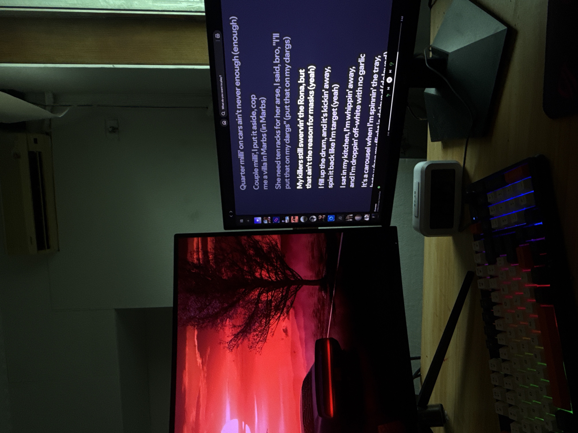 Here is a description of the image:
The image shows a desk setup featuring two computer monitors, a keyboard, and a small electronic device. The top monitor displays the lyrics of a song, with the search query visible in the search bar. The bottom monitor displays a red-toned nature-themed wallpaper, likely a tree and landscape. A mechanical keyboard with RGB lighting is placed to the right of the monitors. A small white device sits on the desk in front of the keyboard. The background is blurred, but elements of a wall and an air conditioning unit can be seen. The photograph seems to be taken indoors, possibly in a home office or gaming room.