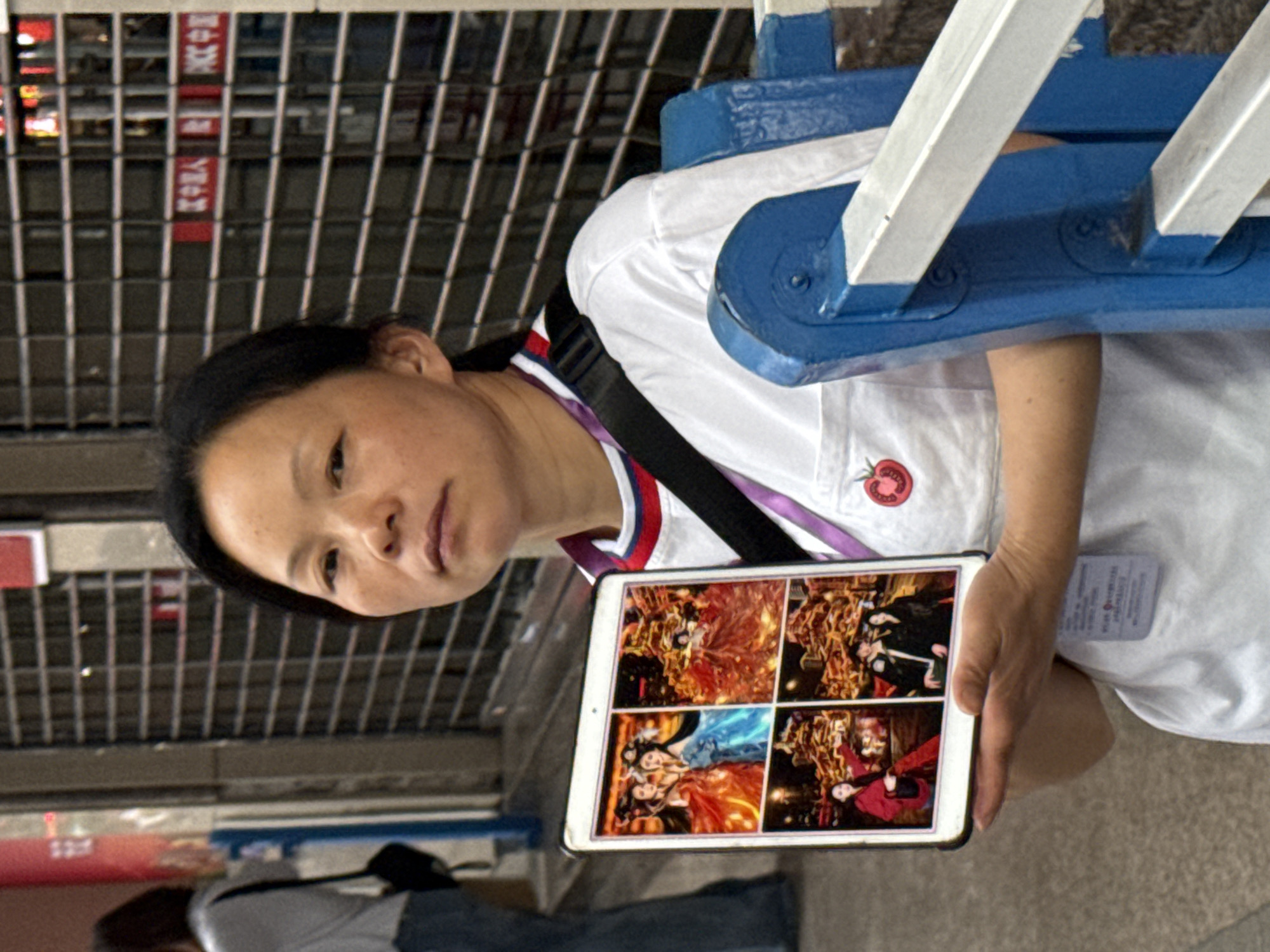 A woman with black hair stands in Yuzhong District, China, holding a tablet displaying four images of costumed performers. She wears a white t-shirt with a tomato emblem and a black shoulder strap. A metal grid and a blue and white structure are visible in the background. Another person, wearing a grey shirt and jeans, is partially visible.