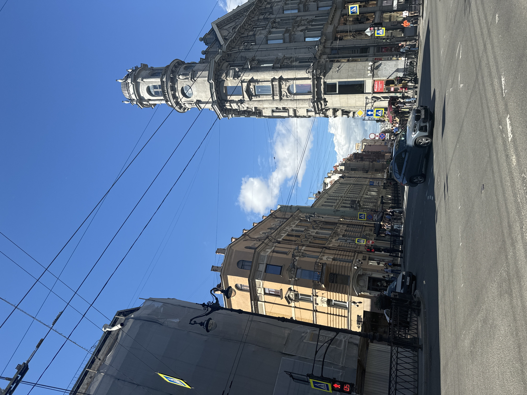 The image depicts a street intersection in Saint Petersburg, Russia, under a clear blue sky with scattered clouds. On the right, a distinctive, ornate grey building with a prominent tower, featuring multiple windows and architectural embellishments, stands at the corner. To the left, a building undergoing renovation is covered in grey tarpaulin, with a traffic light displaying a red signal. Further down the street on the left, a building with a pale yellow facade and numerous windows is visible. The intersection is paved with asphalt and marked with white lines. Several cars are present, including a grey SUV in the foreground and a white sedan in the background. Pedestrians are visible on the sidewalks and at crosswalks. Overhead, a complex network of electrical wires crisscrosses the sky. The overall scene captures a busy urban environment on a sunny day.