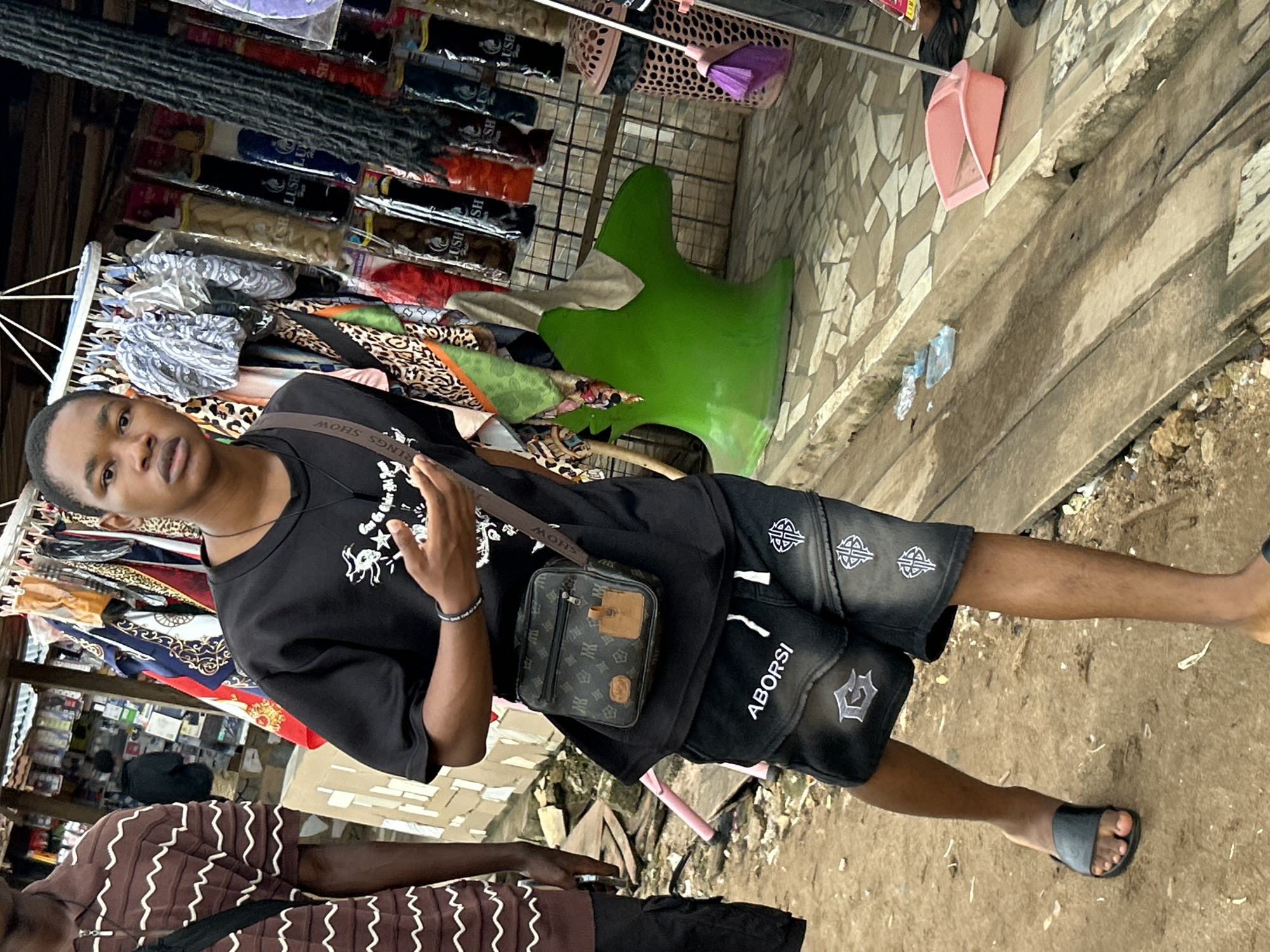 In Ihiagwa, Nigeria, a young Black male stands facing the camera on a dirt and tiled walkway adjacent to a market stall. He is wearing a black t-shirt with a white graphic, black denim shorts with white lettering and embroidered designs, and black slides. A brown and black patterned crossbody bag is slung over his left shoulder. His right hand is held up, palm facing outward, in a gesture. Behind him, the market stall displays various clothing items and hair products. To the right of the stall, a bright green, abstract sculpture is visible, with a beige cloth draped over its top. A pink dustpan and a broom with a purple brush lie on the tiled ground near the sculpture. Another person, partially visible on the left, wears a brown and white horizontally striped polo shirt. The environment suggests an outdoor marketplace setting with goods displayed for sale.