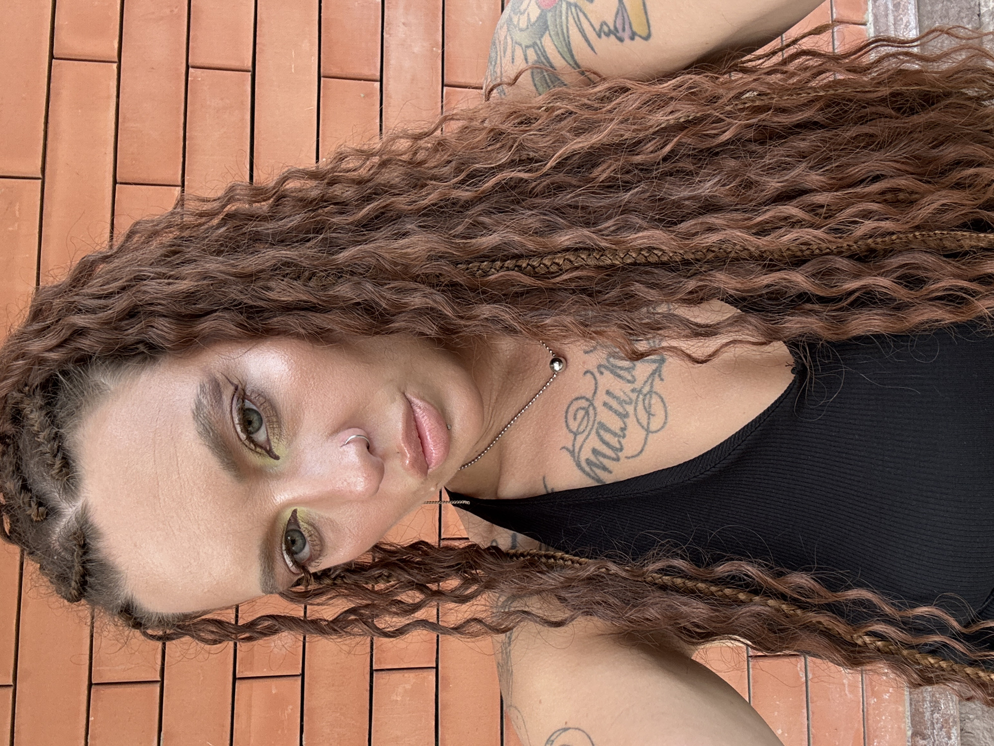 This is a vertical, close-up, selfie-style photograph of a person with long, wavy, auburn hair that is braided in some sections. The person's face is turned towards the camera, with their eyes open and looking slightly upward. They are wearing a black tank top and have several tattoos visible on their chest and arm, including script lettering on the chest. A silver chain with a spherical pendant is visible around their neck. The person is also wearing a silver nose ring. Their makeup includes green eyeshadow.
The background consists of a wall made of reddish-brown bricks laid in a horizontal pattern. The lighting suggests it is daytime, possibly outdoors due to the natural light. The overall mood appears casual and personal. No specific activity or event is depicted beyond posing for the photograph. The location context provided is Grushevskaya, Russia, but there are no specific visual cues in the image to confirm this location.
