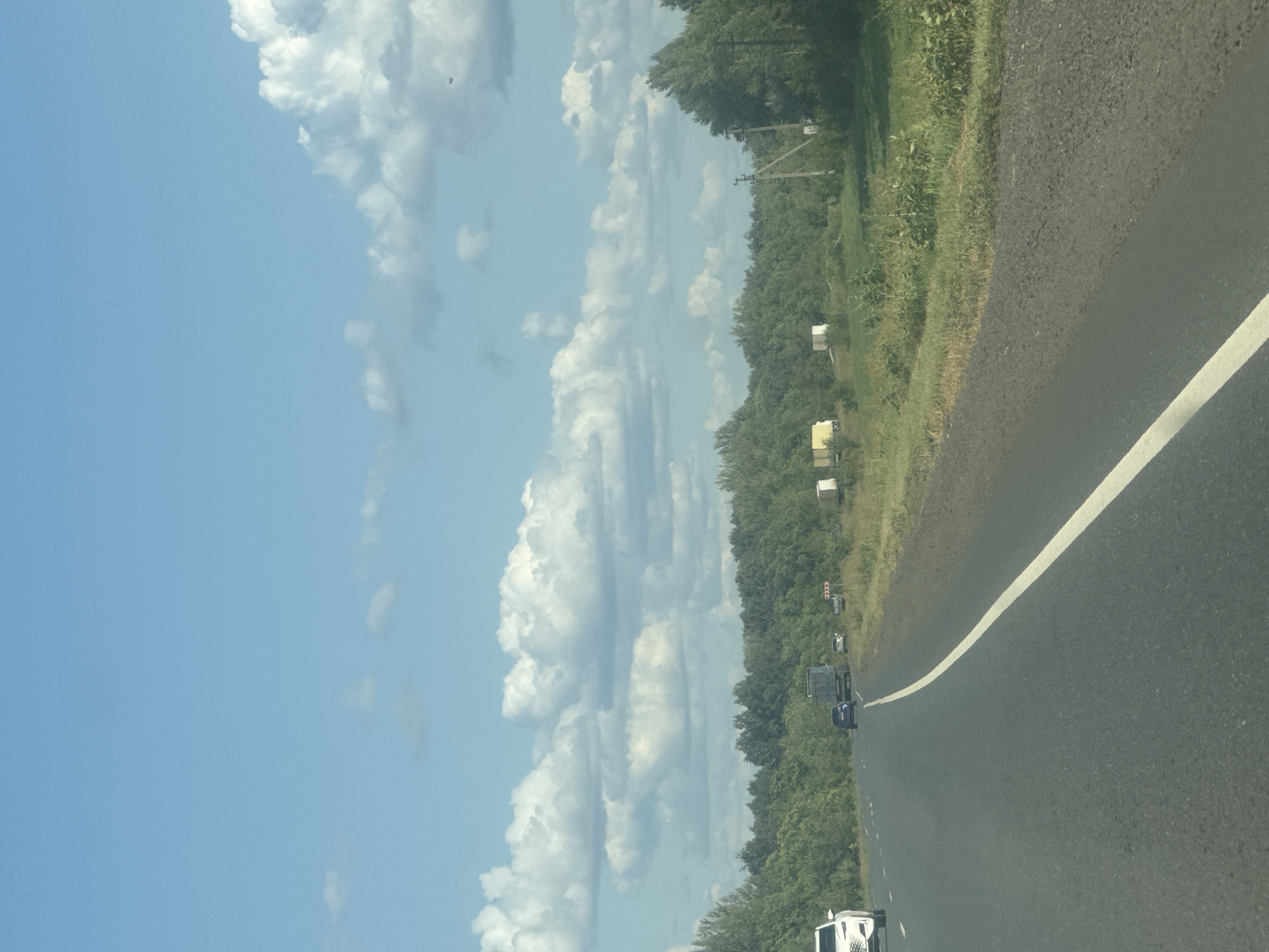 The image depicts a highway scene in an outdoor, rural setting. The perspective is from a moving vehicle, looking forward along the asphalt road which has a white line marking the right lane. The road curves slightly to the left and ahead, several vehicles are visible, including a white SUV on the left, a dark-colored car, and a truck. A dense line of green trees forms a backdrop along the right side of the road, extending into the distance. Beyond the trees, the sky is a clear blue with scattered white, fluffy clouds. The environment appears to be a landscape with vegetation and open sky. There are no people clearly visible in the image. The context provided indicates this scene is located in the city of Novosergiyevka, Russia.