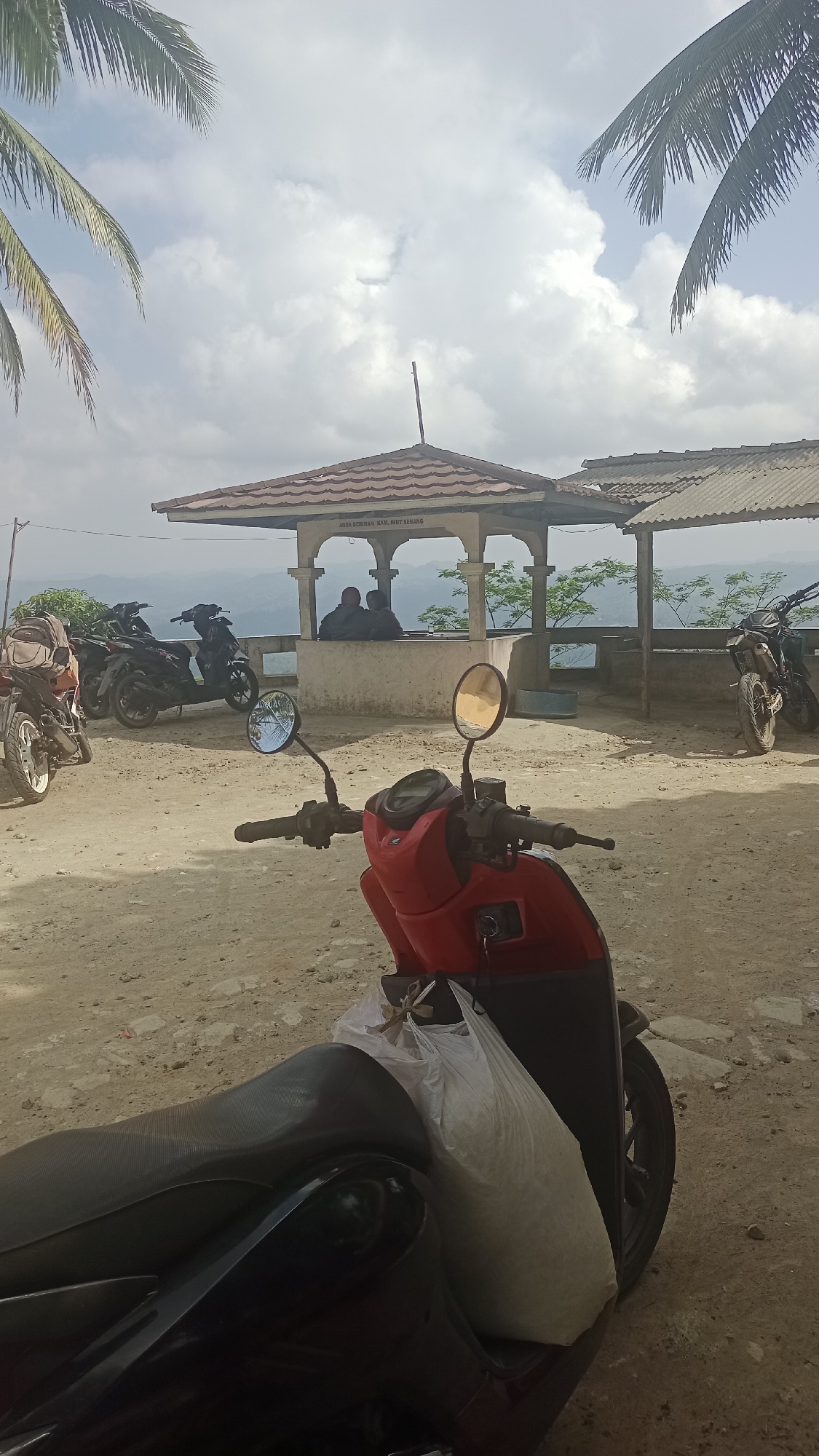 In Karyamandala, Indonesia, a dirt lot is filled with parked motorcycles under a cloudy sky with palm fronds framing the upper portion of the image. A red scooter with a white bag on its seat is in the foreground. Further back, several other motorcycles are parked near an open-air pavilion with two people sitting inside. The pavilion overlooks a valley with rolling hills. The scene appears to be a viewpoint or resting area.