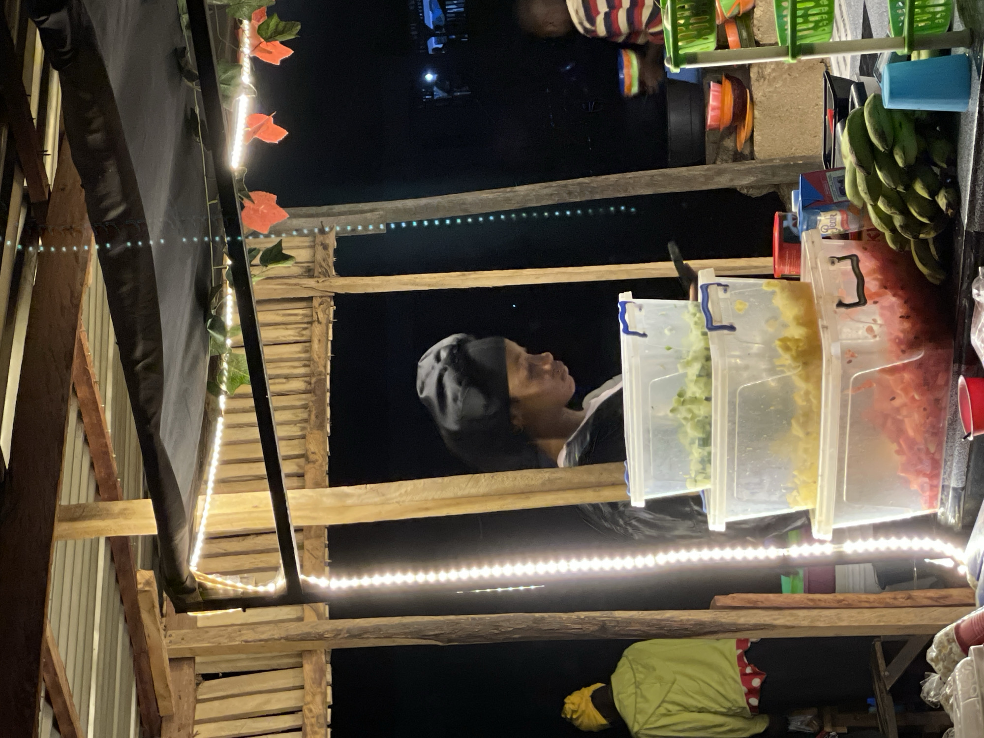 The image depicts an outdoor food stall at night, likely in Durumi, Nigeria. A person wearing a black head covering and a black jacket is standing behind a counter filled with clear plastic containers holding chopped food. The containers are stacked, with the top one holding green chopped food, the middle one holding yellow chopped food, and the bottom one containing red chopped food. To the left, another person wearing a yellow head covering and a bright green jacket is visible. The stall is illuminated by strings of white lights and some artificial orange leaves. In the background, a person in a striped shirt is seen holding a colorful bowl. Various kitchenware, including bowls, baskets, and a bunch of green bananas, are also present on the counter. The environment appears to be a casual, open-air setting.