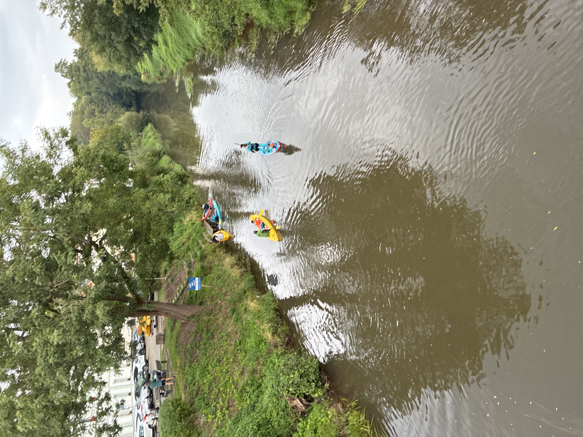 In Kuks, Czechia, a group of people are enjoying a leisurely day on the river. The image captures a view from a bridge looking down on the Elbe River. Several individuals are engaged in water activities. One person is paddleboarding, standing upright on a board and using a paddle. Nearby, two people are in a yellow kayak, paddling downstream. Further up the river, another kayak is visible with two occupants. The riverbanks are lined with lush green grass and trees, with some parked cars visible on the left side of the image, suggesting the proximity of a town or populated area. The sky is overcast, casting a soft, diffused light on the scene.
