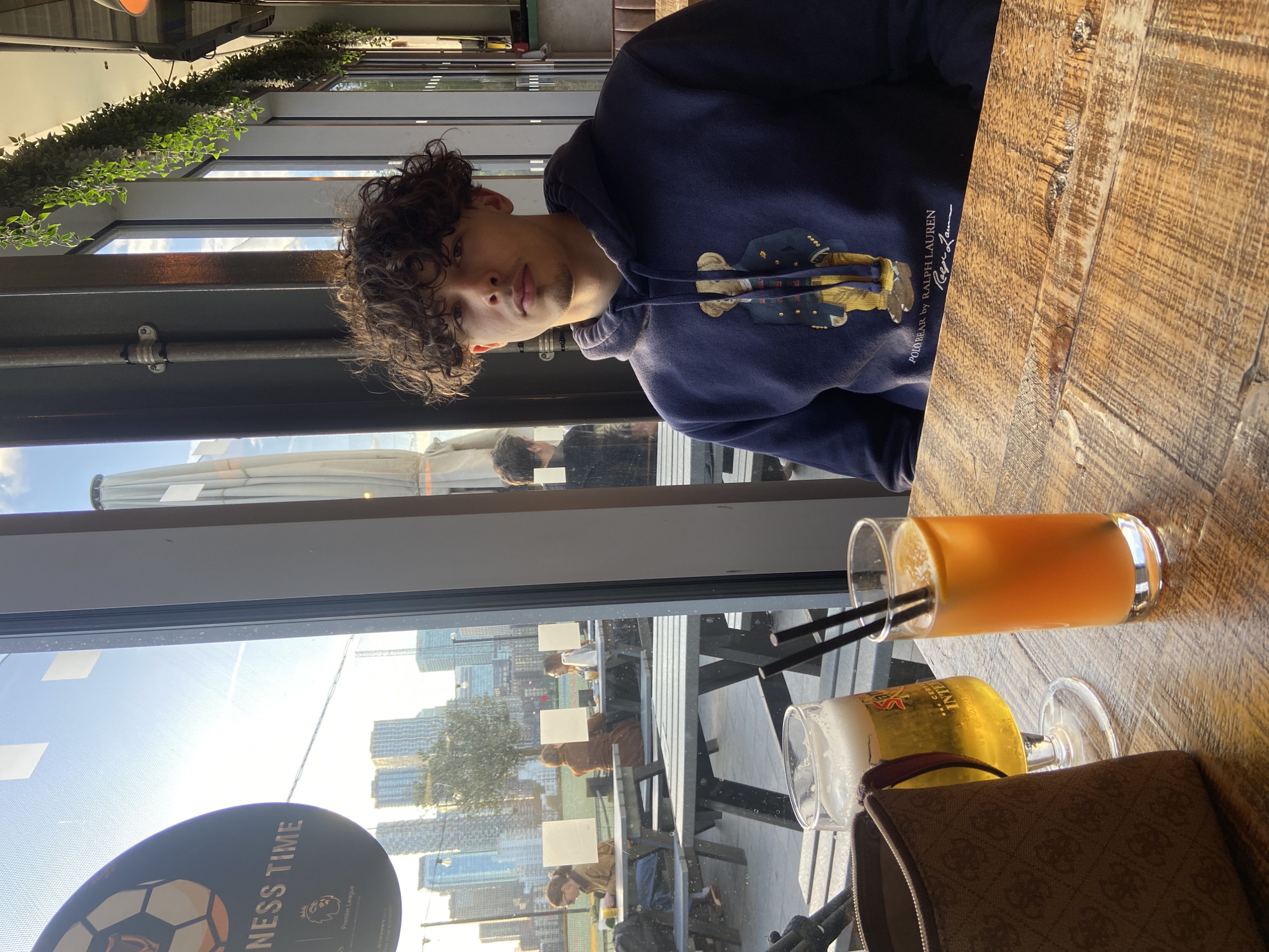 The image, taken in London, UK, captures a young man seated indoors at a wooden table in what appears to be a cafe or restaurant. He has curly dark hair and a light goatee, looking directly at the viewer with a neutral expression. He is wearing a navy blue hooded sweatshirt with a "Polo Bear by Ralph Lauren" design on the front, and the text "POLO BEAR by RALPH LAUREN" along with a signature is visible on the garment.
On the rustic wooden table, there are two drinks: a glass of orange-colored juice with two black straws and a glass of beer, partially obscured by a brown patterned bag. A portion of text, possibly "INTER," is visible on the beer glass.
Behind the man, a large window reveals an outdoor patio area with several people seated at picnic-style tables. Beyond the patio, a modern city skyline with numerous tall buildings is visible under a bright, sunny sky. The strong daylight creates reflections on the window surface. Inside, artificial green foliage decorates an overhead structure.
A circular sign is partially visible through the window, displaying the text "NESS TIME" and the "Premier League" logo, suggesting a football-themed venue or event. The overall setting indicates a relaxed daytime gathering in an urban environment.