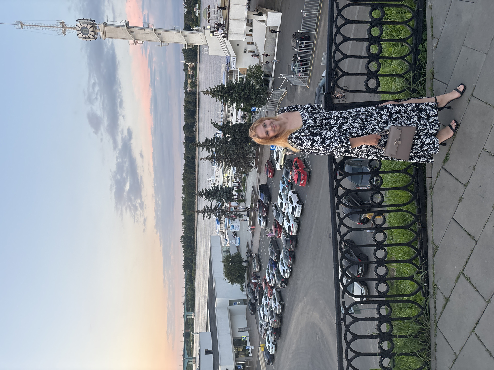 This image depicts a woman standing on a paved walkway with a black metal railing in the foreground. She is wearing a long black and white floral print dress with a thigh-high slit, black strappy high heels, and carrying a light brown handbag. Behind her, a river flows, with a bridge visible in the distance. A large parking lot filled with cars is situated next to a white building with a flat roof. To the right, a tall white tower with a clock face stands prominently. Trees line the riverbank, and several boats are docked. The sky exhibits the soft hues of either sunrise or sunset with clouds. The scene is likely located in Yaroslavl, Russia, specifically along the Volga River embankment, near the Strelka (Arrow) peninsula, given the presence of the iconic tower and the general cityscape.
