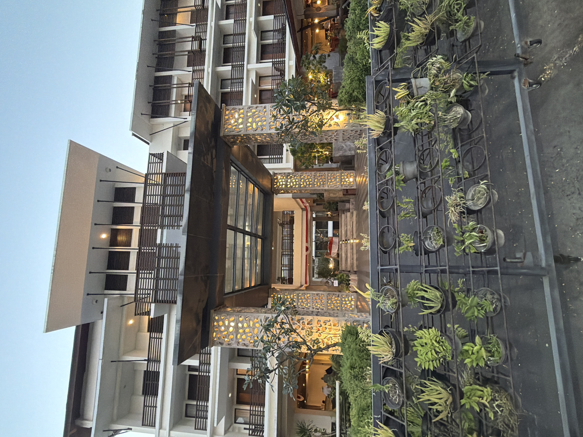 The image shows the exterior of a modern hotel in Kuta, Indonesia, at dusk. The hotel has multiple floors with balconies and is illuminated by warm lighting. In the foreground, there is a tiered metal structure holding numerous potted plants, primarily succulents and small green plants. A walkway leads to the hotel's entrance, which features a covered portico with a glass roof and decorative illuminated pillars. The sky above is a pale blue, indicating the transition from day to night. No people are clearly visible in the scene.