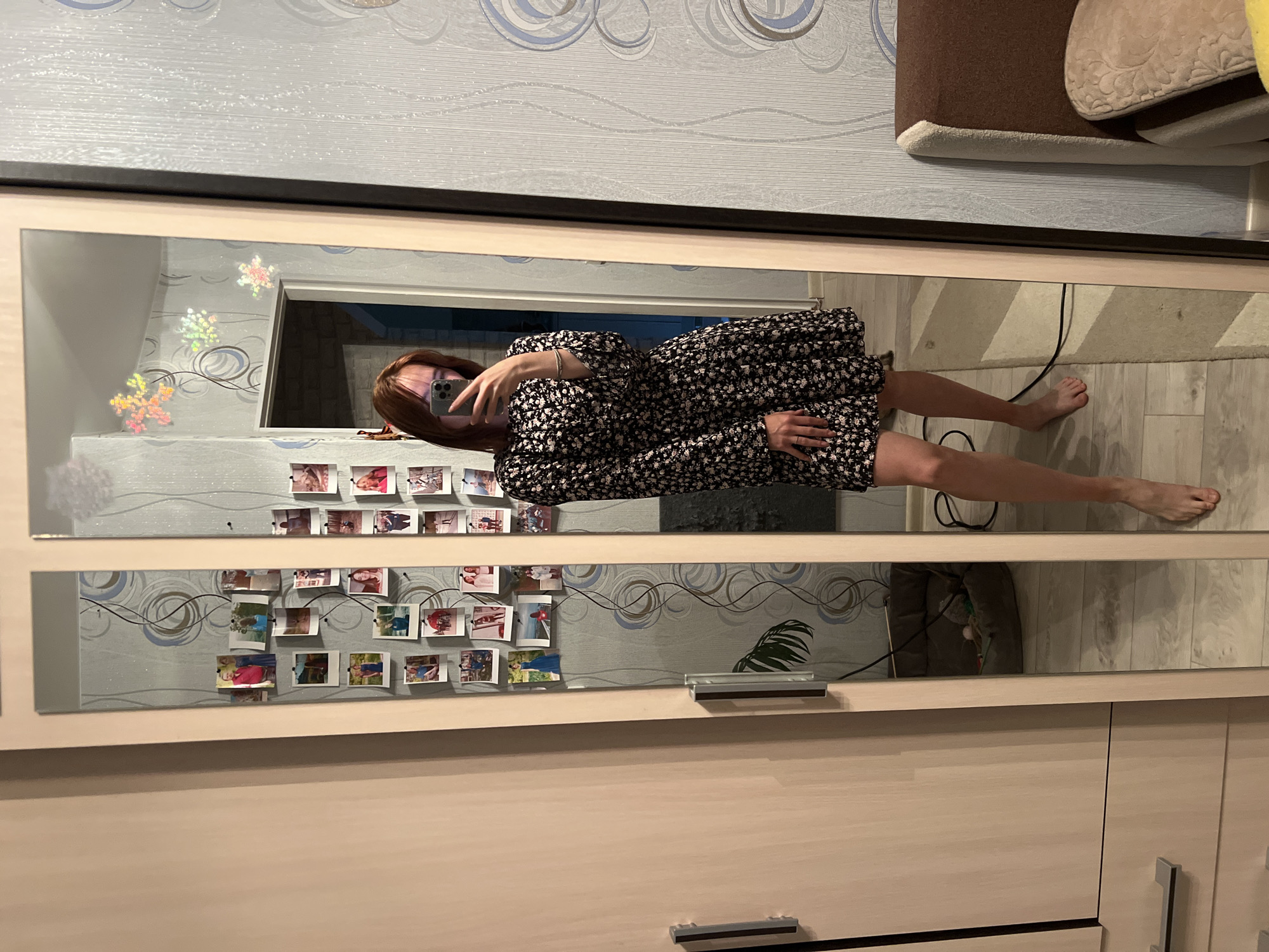 In a brightly lit room, a young woman with auburn hair is captured taking a full-length selfie in a floor-to-ceiling mirror. She is wearing a dark, floral-patterned dress with long sleeves and a flared skirt. Her bare feet are visible, as is a thin bracelet on her left wrist. The mirror reflects a wall adorned with numerous small photographs and some holographic star decorations. To the left of the mirror is a wardrobe with light-colored doors and silver handles. The floor appears to be laminate wood. In the reflection, a pet bed can be seen in the background, along with a doorway leading to another room. The overall environment suggests a residential setting. The image was taken indoors, possibly in an apartment in Chaikovsky, Russia, given the provided metadata.