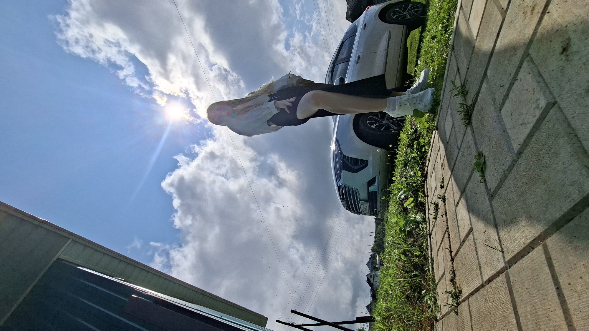 A young woman stands in front of a light-colored SUV in what appears to be a rural setting near the village of Bolshoy Bukor, Russia. The woman is seen from the waist down, wearing a white long-sleeved shirt and a dark skirt, with white sneakers. She is positioned in front of the driver's side of the vehicle. The SUV is parked on a grassy area, with houses and trees visible in the background. The sky is partly cloudy with bright sunlight, creating a lens flare effect. In the foreground, there is a paved walkway made of rectangular concrete pavers, with grass growing in the cracks. The overall scene suggests an outdoor recreational or travel moment.