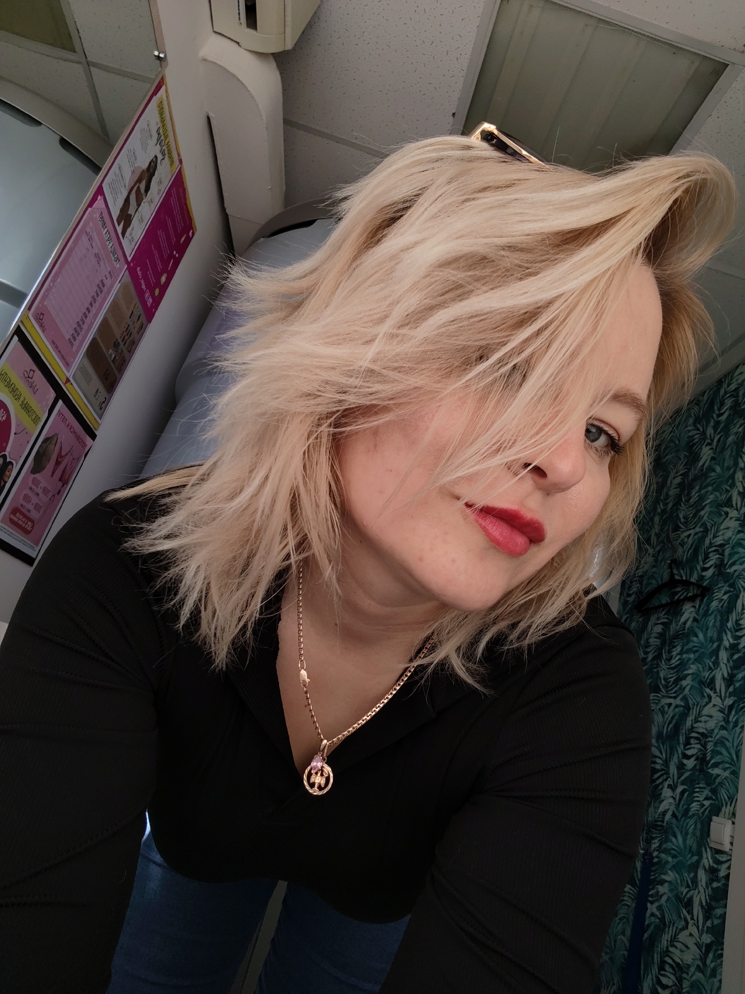 A woman with blond hair is pictured indoors. She has fair skin, blue eyes, and is wearing red lipstick. Her hair is styled with strands falling across her face. She is wearing a black long-sleeved shirt and a gold necklace with a circular pendant. A portion of blue denim can be seen below the shirt. Behind her is a wall with a dark green patterned covering. Above her head is a white object that appears to be an air conditioning unit. To her left is a board with advertisements written in a non-English script. Location details suggest Izhevsk, Russia.