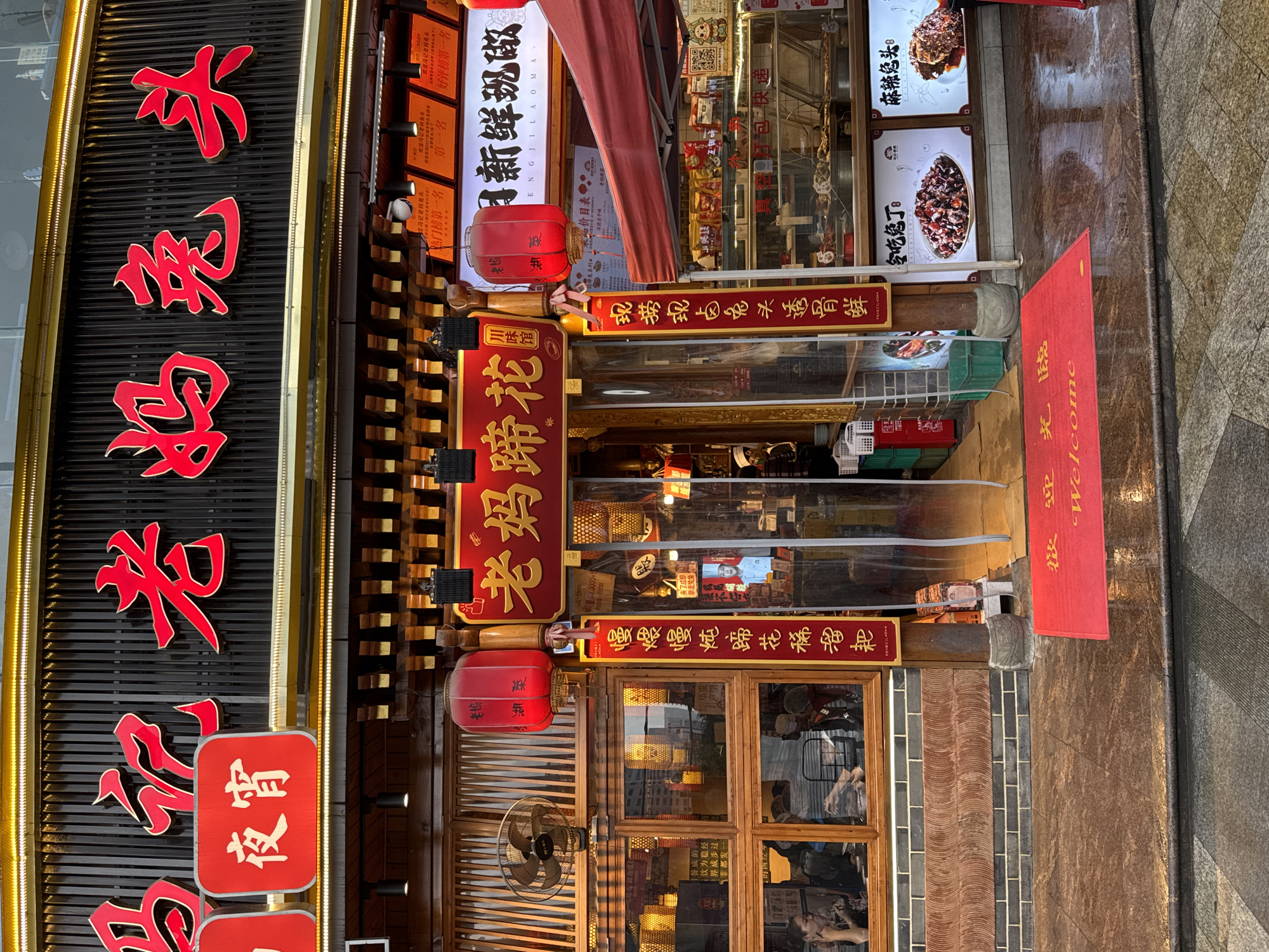 The image shows the front of a Chinese restaurant or shop, located in Jinjiang District, China. The sign on the left reads "Ma Ji Lao Ma Tu Tou" (马记老妈兔头). Other signs indicate the restaurant sells rabbit head (兔头) and pig trotters (蹄花). One sign says "Fengji Lao Ma Tu Tou" (冯记老妈兔头). A red welcome mat is at the entrance. The entrance is partially covered with plastic curtains. There are lanterns and lights illuminating the shop. The shop's windows show reflections of the surrounding environment. A rotating fan is visible near the door. Food items are advertised on posters. The street is paved with stone.