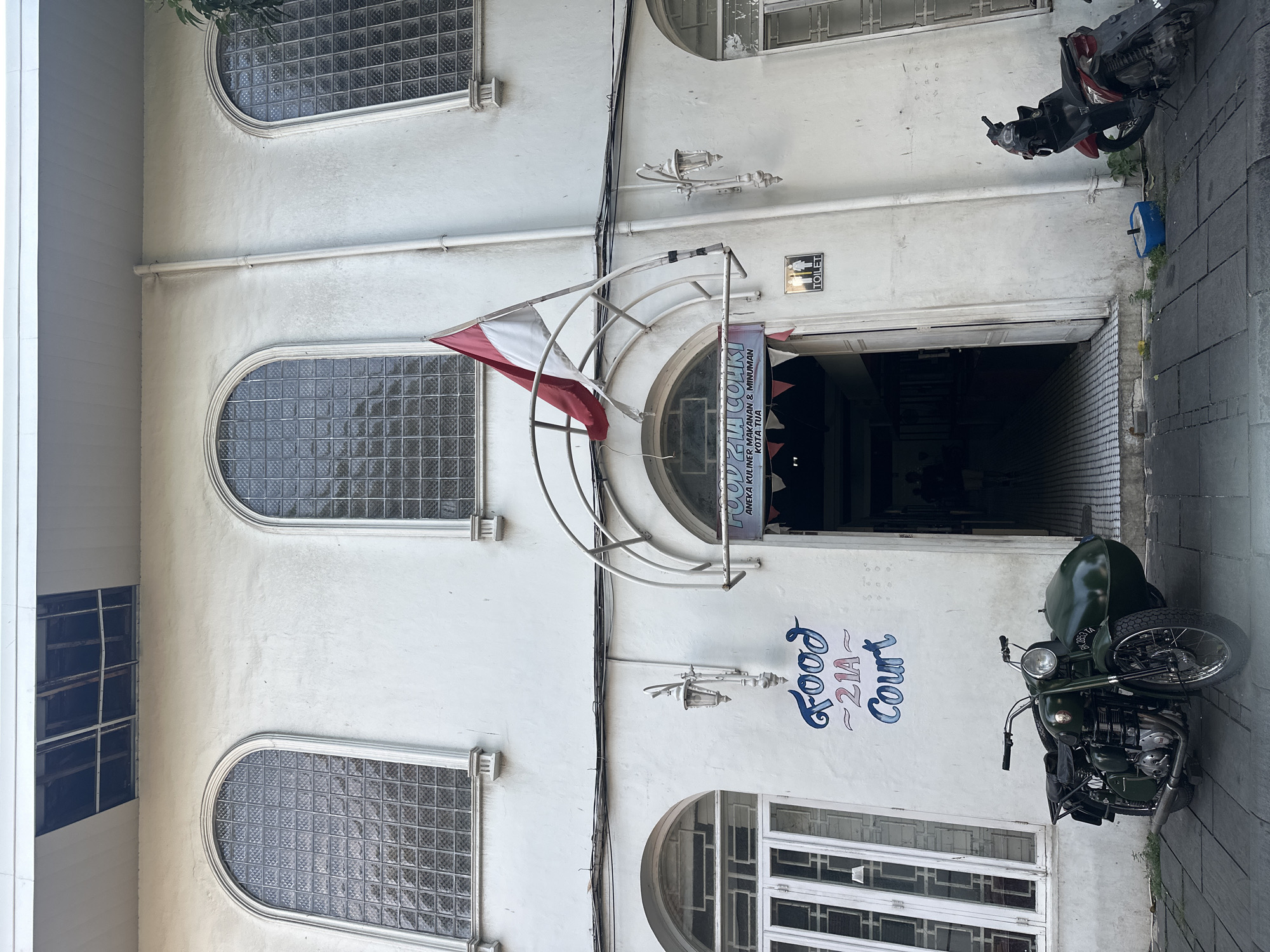 This vertical photograph shows a building with rounded-top windows, a sign that reads "FOOD 21A COURT," and two parked motorcycles. The building appears to be a food court, indicated by the sign above the entrance that reads "FOOD 21A COЧКІ, ANEKA KULINER MAKANAN & MINUMAN KOTA TUA." A white and red flag is affixed to a semi-circular structure above the entrance. A motorcycle is parked on the right side of the entrance, and another is slightly further away, closer to the top of the image. The walls are white, and a street or sidewalk is visible to the right of the building. A "TOILET" sign is next to the entrance. The photo suggests the location is in Kota Tua (Old Town) in West Jakarta, Indonesia, as indicated on the sign.