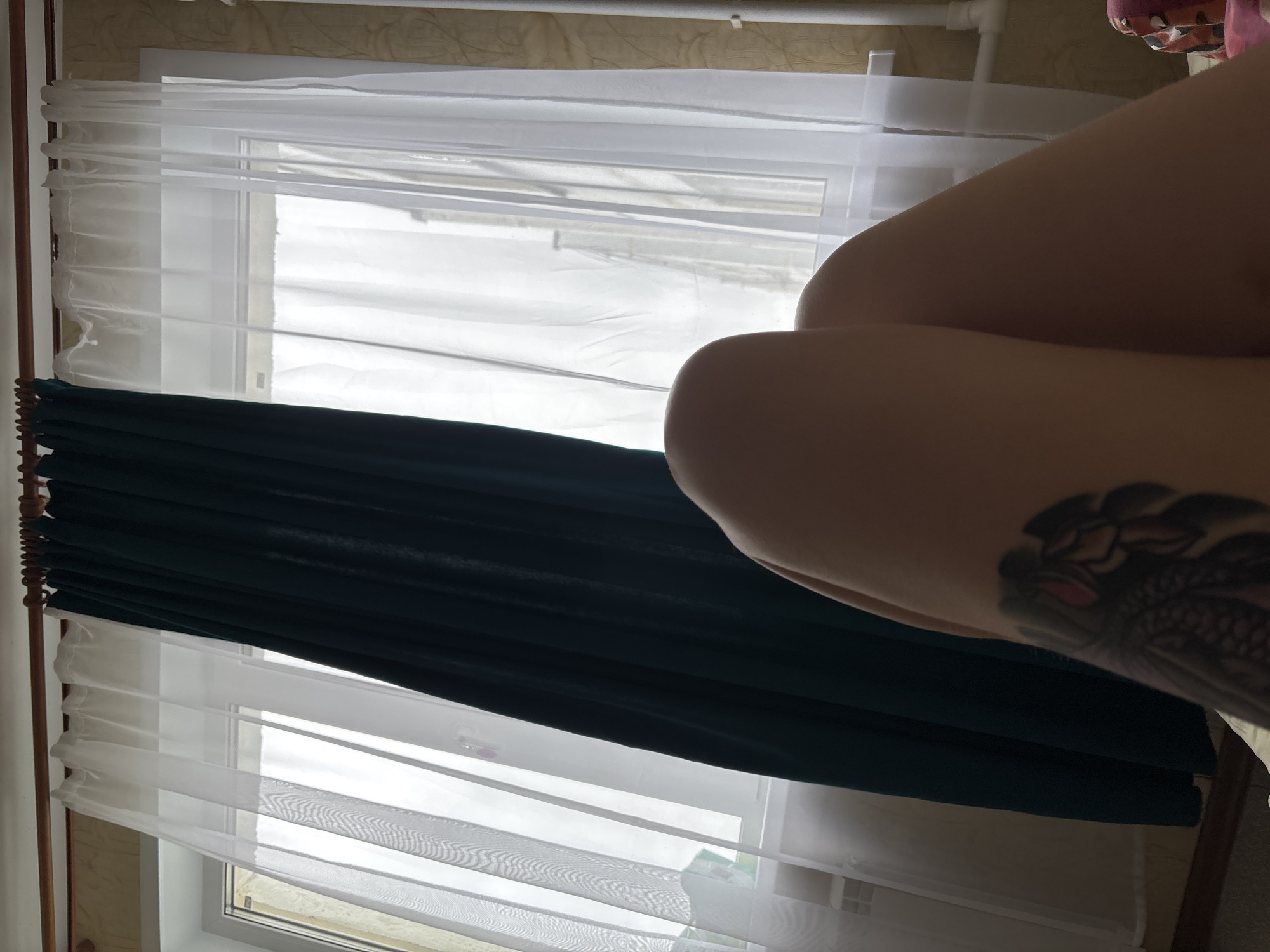 The image is a close-up, low-angle shot of a person's legs and knees, partially obscuring a window with sheer white curtains and a dark teal curtain drawn to the side. The person has a tattoo on their left thigh, featuring a dark, stylized bird or mythical creature with pink accents. The background shows a wall with a textured wallpaper pattern and a white radiator pipe. The scene appears to be indoors, possibly a bedroom, with natural light filtering through the window. There is no identifiable indication of the location being in Naberezhnye Chelny, Russia.