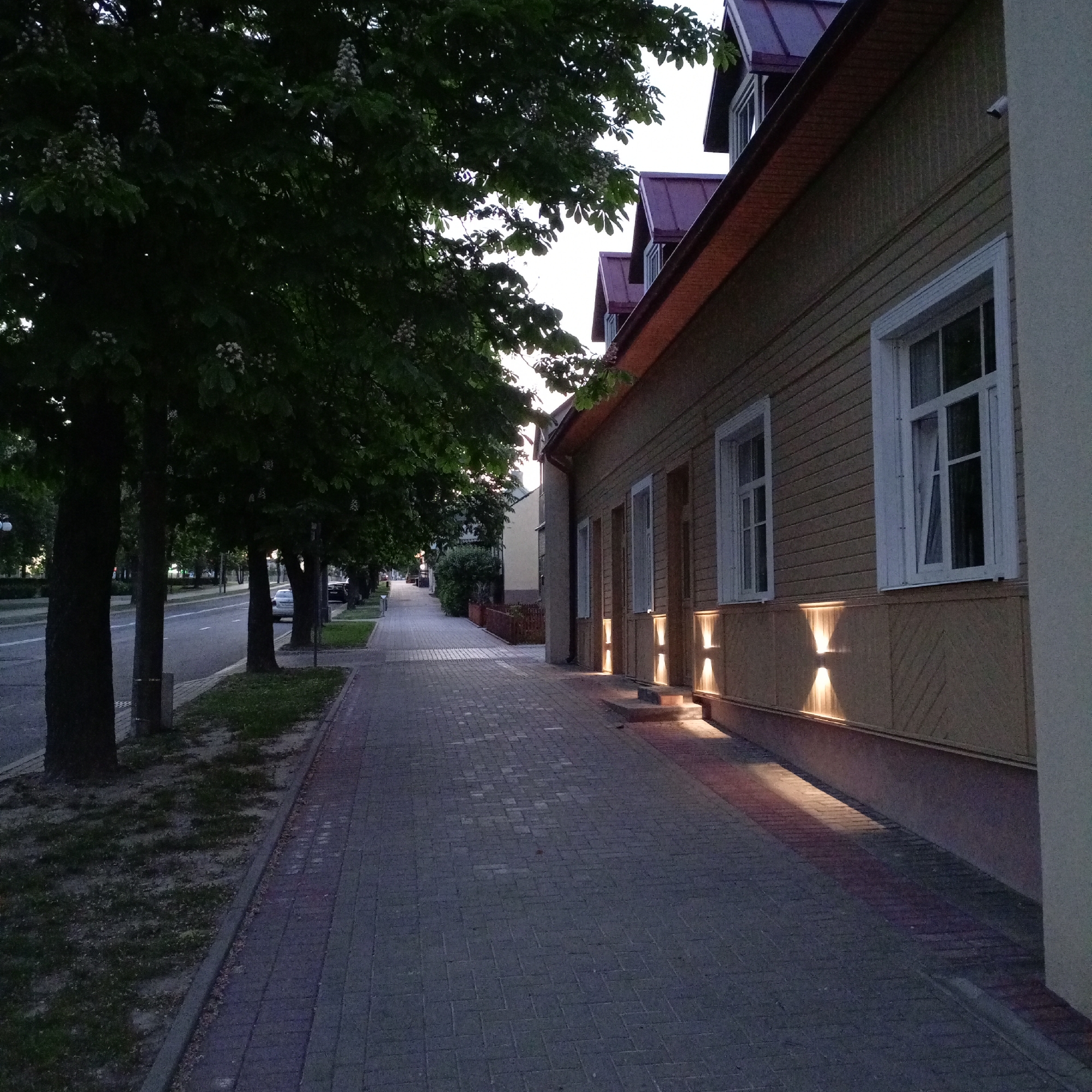 The image depicts a sidewalk in an urban setting, possibly in the city of Unknown, Unknown. The sidewalk is paved with grey rectangular blocks. To the left of the sidewalk are trees and grass, with a road running parallel to them. Cars are parked along the road. To the right of the sidewalk is a light brown wooden building with white-framed windows. Exterior wall-mounted light fixtures are present on the building's wall, emitting light onto the sidewalk. The image seems to have been taken during dusk or dawn.