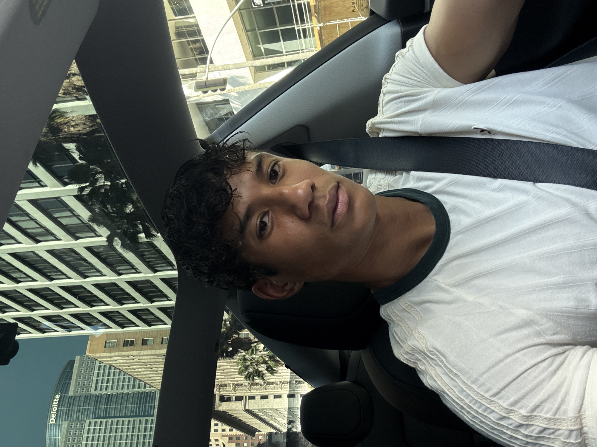 A person is wearing a white t-shirt and a seatbelt in the front seat of a car. The reflection of the Deloitte building can be seen in the windshield. The reflection of the Deloitte building indicates the location as downtown Los Angeles, California, United States of America.