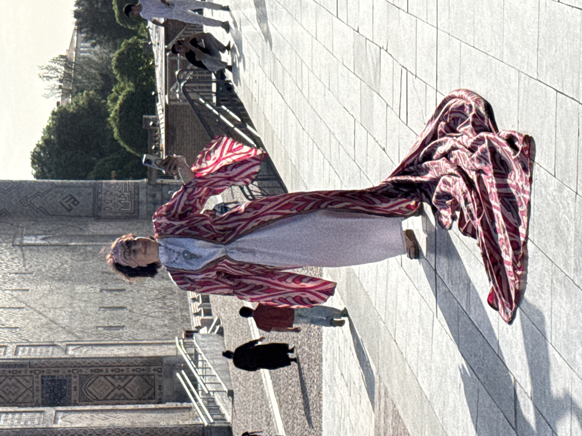 The image depicts a woman in Samarkand City, Uzbekistan, seemingly taking a photo with her phone in front of an ornate building. She is wearing a long, patterned robe or dress, primarily in shades of red and white, with voluminous sleeves and a gathered train-like extension. A white underdress or garment is visible beneath the robe. She also wears a head covering or headband.
The building behind her features intricate geometric patterns and designs, characteristic of Central Asian architecture. The environment suggests a public space or plaza, with a paved surface and other people visible in the background. Several other people are walking on the paved ground. The overall impression is that the woman is a tourist or resident engaging with the cultural heritage of the location.