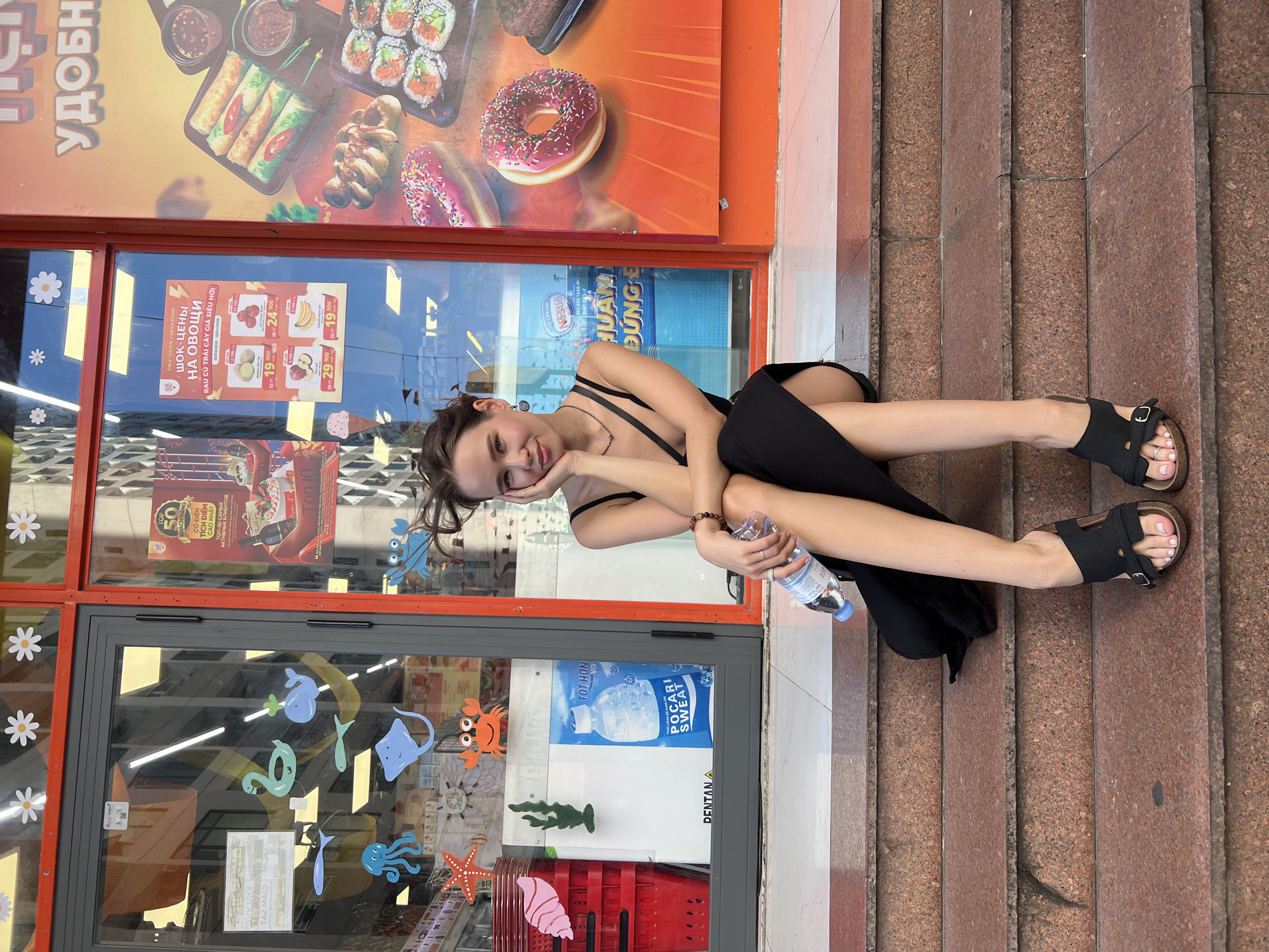 A young woman is captured in a relaxed pose, sitting on reddish-brown stone steps outside a store in Bac Nha Trang Ward, Vietnam. She is positioned on her side, leaning on one hand with a gentle smile directed towards the camera, holding a plastic bottle of water in her other hand. She wears a black sleeveless top or dress and black buckled sandals, revealing white painted toenails.
The background consists of the store's entrance, featuring large glass windows and doors framed in red. Above the entrance, a large advertising sign displays appetizing images of sushi, spring rolls, donuts, and other food items. Through the windows, various promotional posters are visible. One advertises "SALE SHOCK FRESH FOOD" with "ШОК-ЦЕНЫ НА ОВОЩИ" (Shock prices on vegetables) and "RAU CỦ TRÁI CÂY GIÁ SIÊU HỜI" (Super cheap prices for fruits and vegetables) in both Russian and Vietnamese, detailing prices for items like potatoes and bananas. Another poster mentions "POCARI SWEAT" and "Nestle" ice cream, with phrases like "TỐT HƠN VỚI" (Better with). Further signs refer to "TOP 50 KHÁCH HÀNG CÓ ĐIỂM TÍCH ĐIỂM CAO NHẤT" (Top 50 customers with highest loyalty points) and "ТОП-50 САМЫХ АКТИВНЫХ КЛИЕНТОВ" (Top 50 most active clients). The glass doors are decorated with colorful marine-themed stickers including crabs, fish, an octopus, a starfish, and a whale, along with white daisy stickers on a yellow section of the frame. A red shopping basket is partly visible inside the store.
The scene takes place during the daytime, under natural light. The presence of both Russian and Vietnamese text on store signs confirms the specified location context. The woman's expression conveys a cheerful and casual mood.