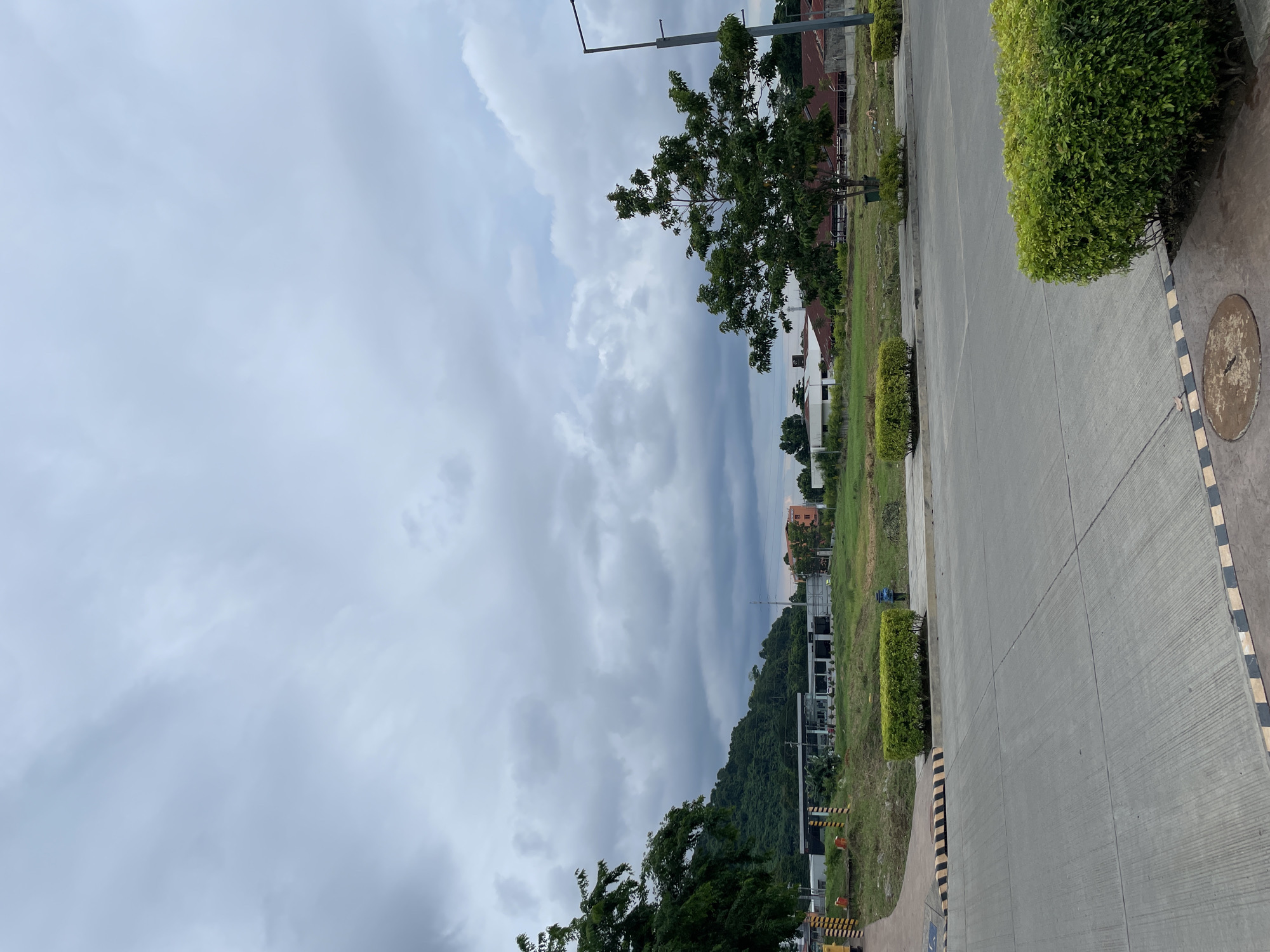 The image depicts a scene in Davao City, Philippines, under a cloudy sky. In the foreground, a paved road with black and yellow striped safety markings is visible. Several manicured green hedges are strategically placed along the edges of the road and sidewalk. Beyond the road, a grassy field stretches out, dotted with more hedges and a few trees. In the mid-ground, a low-rise building with a white facade and dark-framed windows is situated, possibly a commercial establishment or part of a complex. Further in the background, a lush green mountainside dominates the landscape, its slopes covered in dense vegetation. Several smaller buildings with reddish-brown roofs can be seen at the base of the mountain. A street lamp stands on the right side of the image. The overall impression is of an urban fringe area transitioning into a natural landscape. There are no people visible in the image.