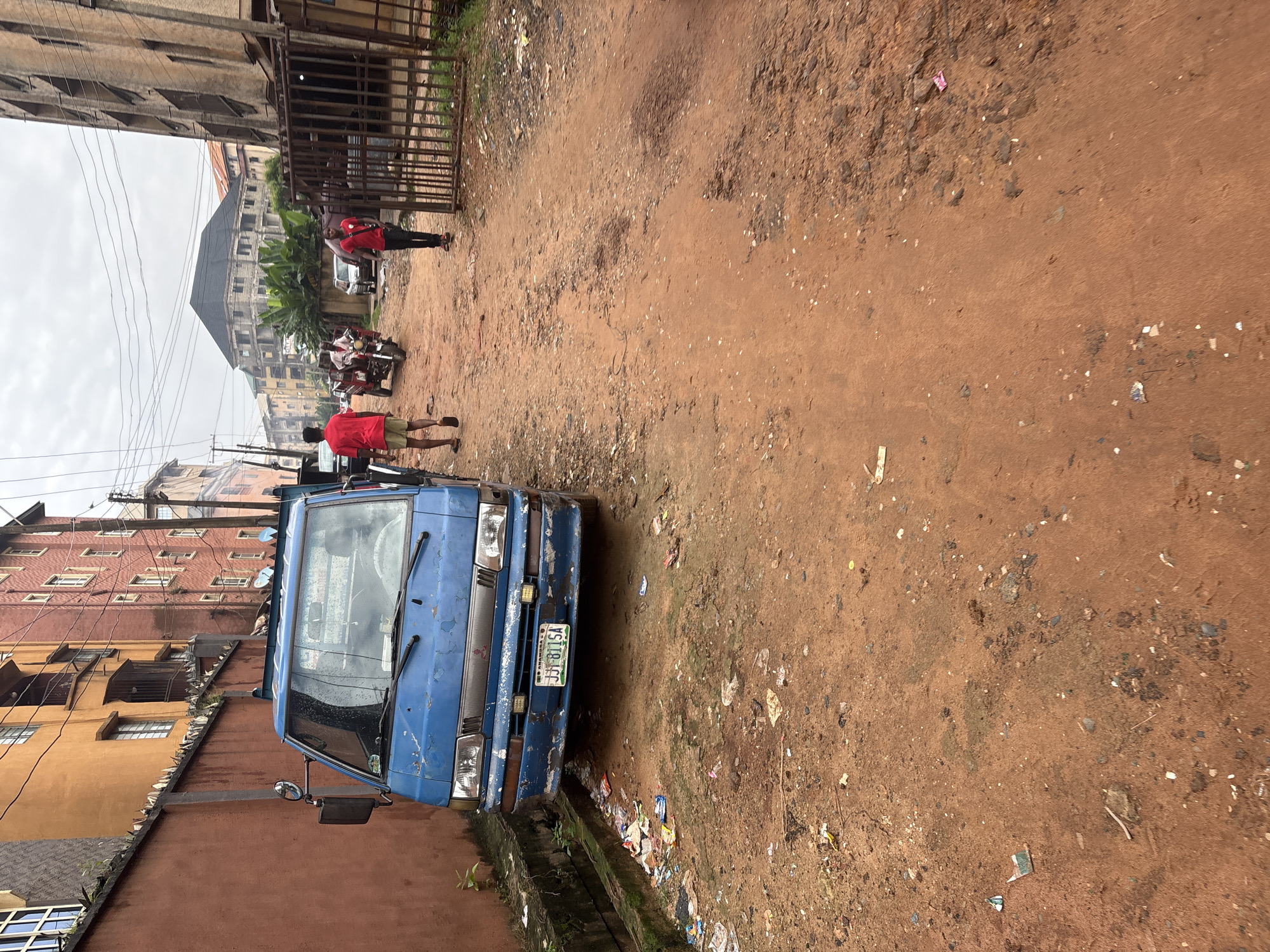 A dusty, reddish-brown dirt road cuts through a residential area in Onitsha, Nigeria. The road is littered with various pieces of trash, including plastic bags, wrappers, and what appears to be a small booklet. On the left side of the road, a blue, slightly weathered delivery van is parked. Its license plate reads "JJJ 811 SA". Adjacent to the van is a tall, orange-colored building with multiple windows and a balcony, bordered by a spiked concrete wall. Further down the road, two individuals wearing red shirts are walking. One is walking away from the camera, and the other is walking towards it. In the background, a motorcycle with a rider is visible, along with several buildings of varying heights and architectural styles, some appearing to be under construction. Power lines crisscross the sky above. On the right side of the road, a metal gate stands open, revealing more of the urban environment beyond.