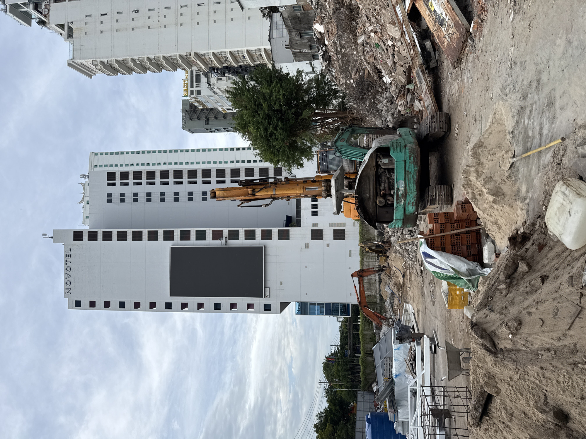 In Nha Trang, Vietnam, a construction site is depicted with a large excavator in the foreground, its engine exposed. Next to it, a pile of bricks and a wheelbarrow suggest ongoing work. Behind the excavator, a vast pile of rubble indicates demolition. In the background, the Novotel Nha Trang hotel stands tall, a white building with a large dark screen on its facade. Other modern buildings and construction materials are also visible. The sky is overcast, with scattered clouds.