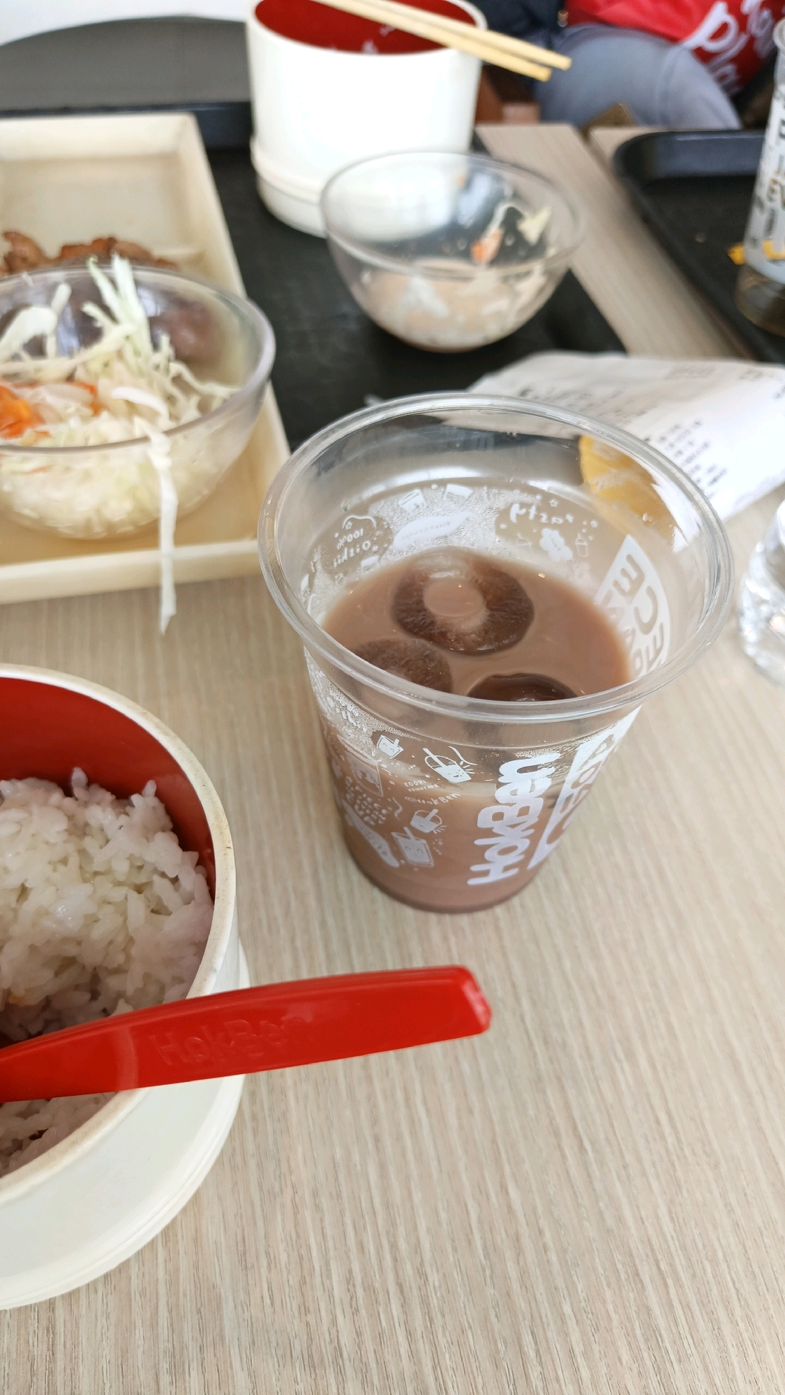 The image displays a close-up view of a meal set on a light-colored wooden table, likely in a dining establishment in Karawang, Indonesia. In the foreground, a white circular container with a red rim holds a portion of white rice. A bright red plastic utensil, possibly a spoon or spork, rests on top of the rice container. To the right, a clear plastic cup contains a light brown beverage with ice cubes. The cup is adorned with a logo and decorative patterns, suggesting it is a branded drink. In the background, several empty clear bowls are visible, along with chopsticks resting on a white cylindrical container with a red interior, hinting at a previous meal or other dishes. A dark tray is also present, along with glimpses of a person's lower body wearing gray pants in the far background. The overall scene depicts a partially consumed meal.