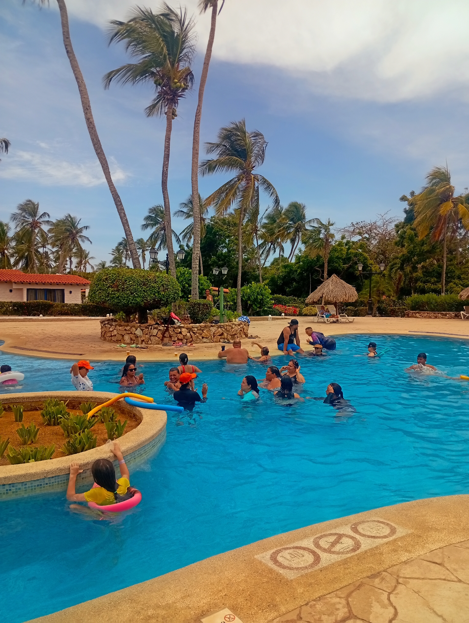 The image shows a swimming pool area at a resort or hotel, likely in a tropical or subtropical location. Many people are in the pool, engaging in various activities: swimming, wading, and socializing. Several palm trees are visible in the background, suggesting a tropical setting. A building with a red-tiled roof is present in the background. The pool has a light blue color and is surrounded by a tan or beige-colored deck. The sky is blue with some clouds. Based on the prompt, the location is Playa El Agua, Venezuela.