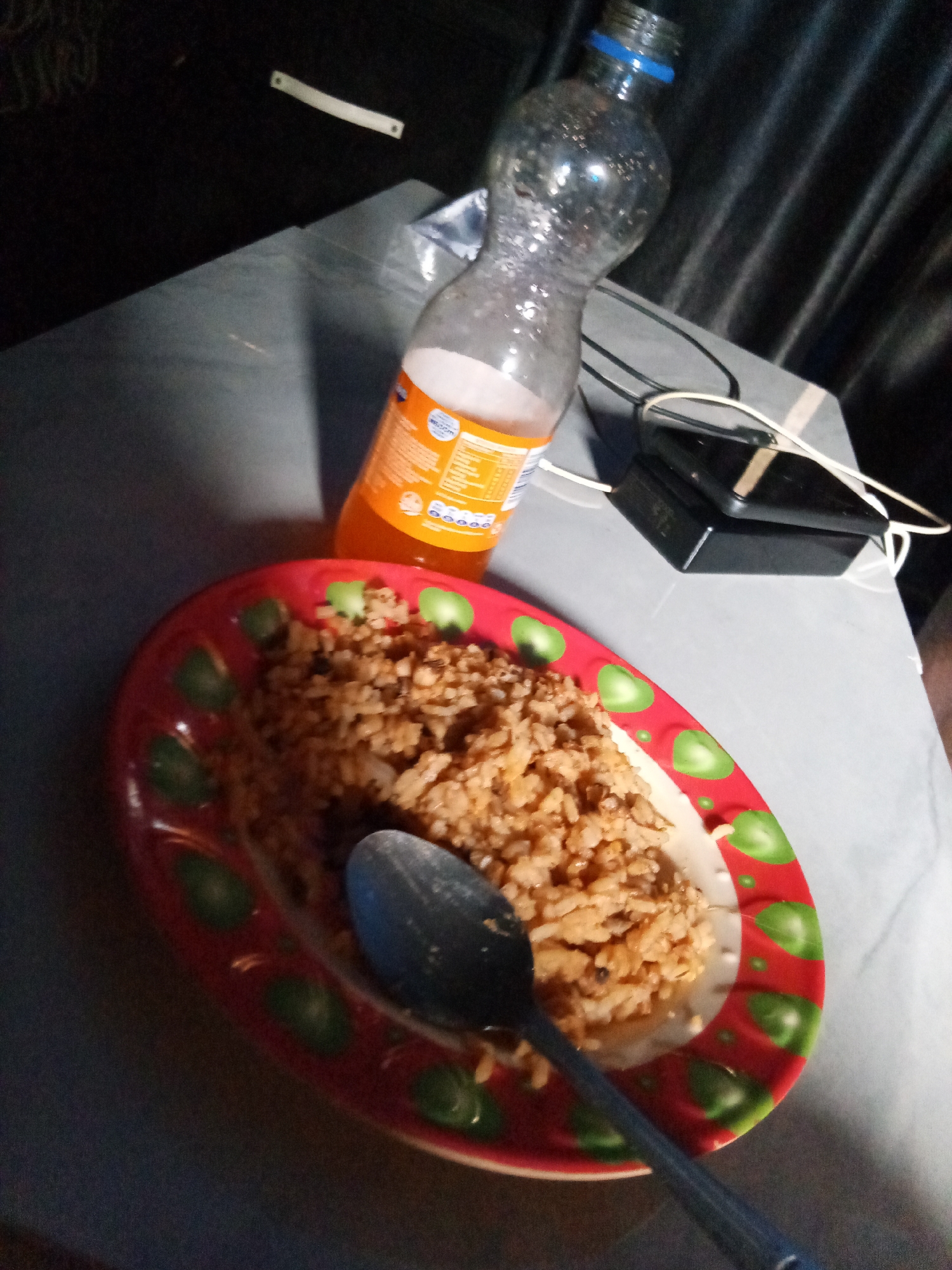 The image depicts a plate of fried rice on a red and green decorated plate with a metal spoon resting on top, accompanied by a bottle of orange soda. A small electronic device and a cable are visible near the soda bottle on a gray surface, which may be a table. The location is unknown.