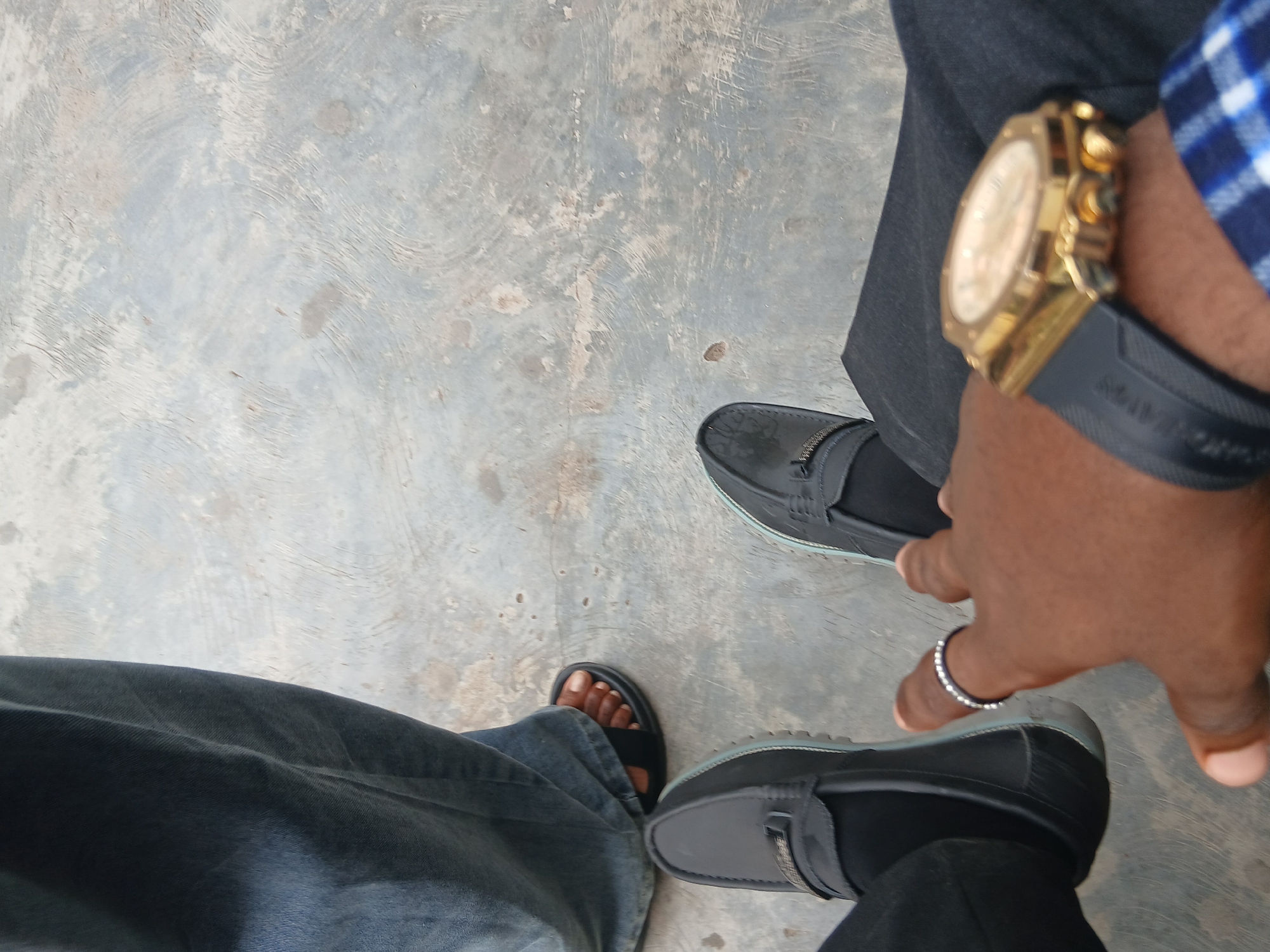 The image shows a ground-level view of two individuals standing on a concrete surface in Lagos, Nigeria. One person is wearing dark gray dress pants, black loafers, a gold wristwatch with a black strap, and a ring on their finger. The other person is wearing blue denim jeans and black sandals, revealing a single foot with exposed toes. The background consists of a simple, gray concrete surface.