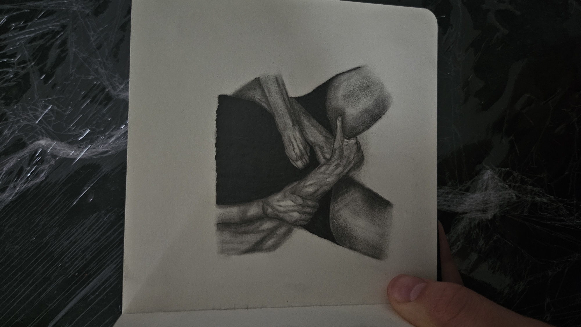 A close-up, overhead shot showcases a sketch of a nude figure in a sketchbook. The sketch, rendered in charcoal, depicts the lower torso and legs of a person, with hands obscuring the groin area. The figure is positioned centrally within the sketchbook, against a plain background. The sketchbook itself is open, with the right page edge visible. The surrounding area is out of focus, with a black, textured surface, possibly a tablecloth or a matte black surface, visible behind the sketchbook. A hand is visible on the left side of the frame, holding the sketchbook. The lighting is soft, highlighting the details of the charcoal drawing.