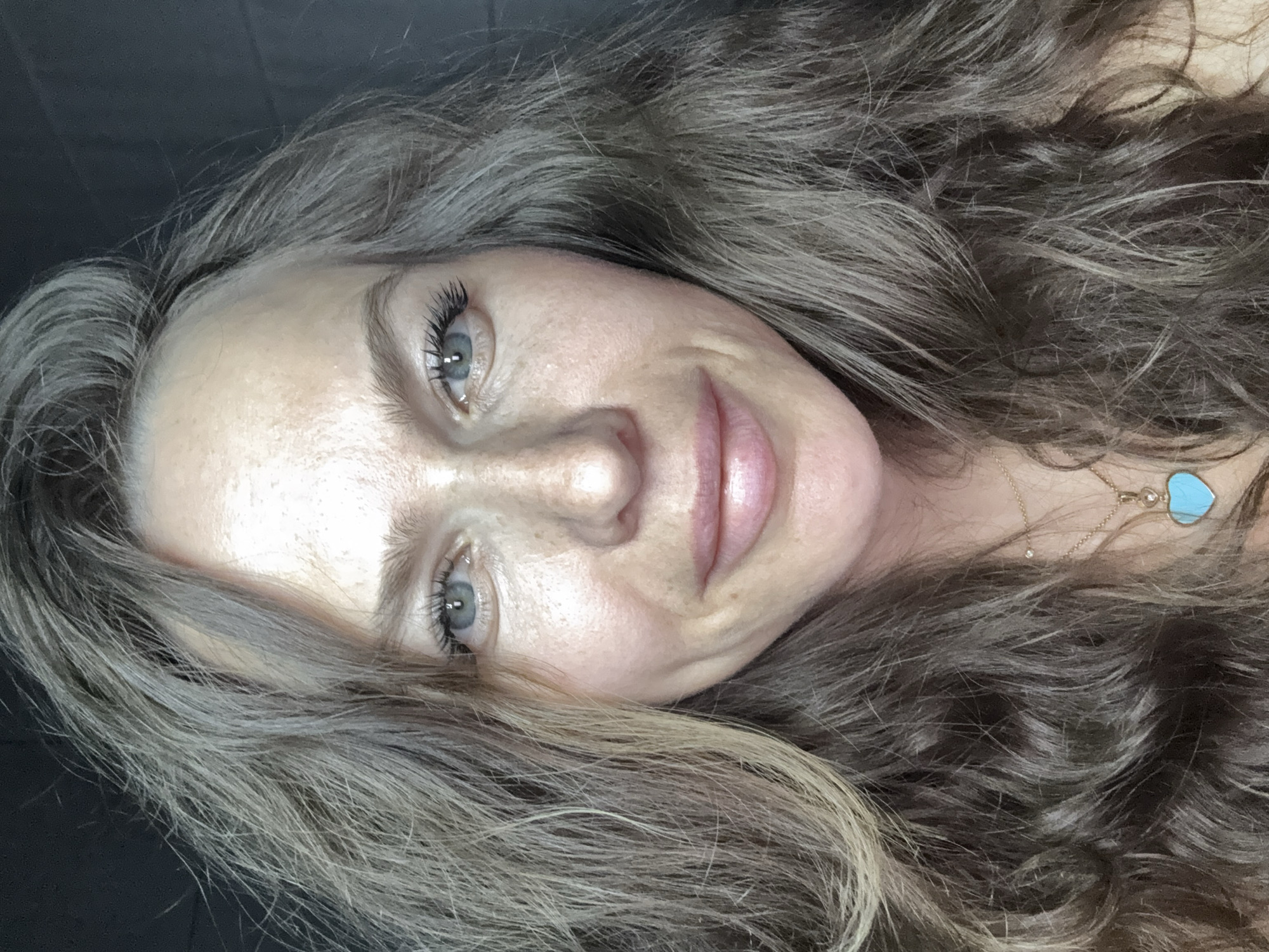 A woman is facing the camera in a close-up selfie. She has fair skin, light blue eyes, and long, wavy hair with light brown and blonde highlights. Her expression is soft, with a slight smile. She is wearing a delicate gold necklace with a turquoise heart pendant. The background is dark and indistinct. There is no indication of the location being Khimki, Russia.