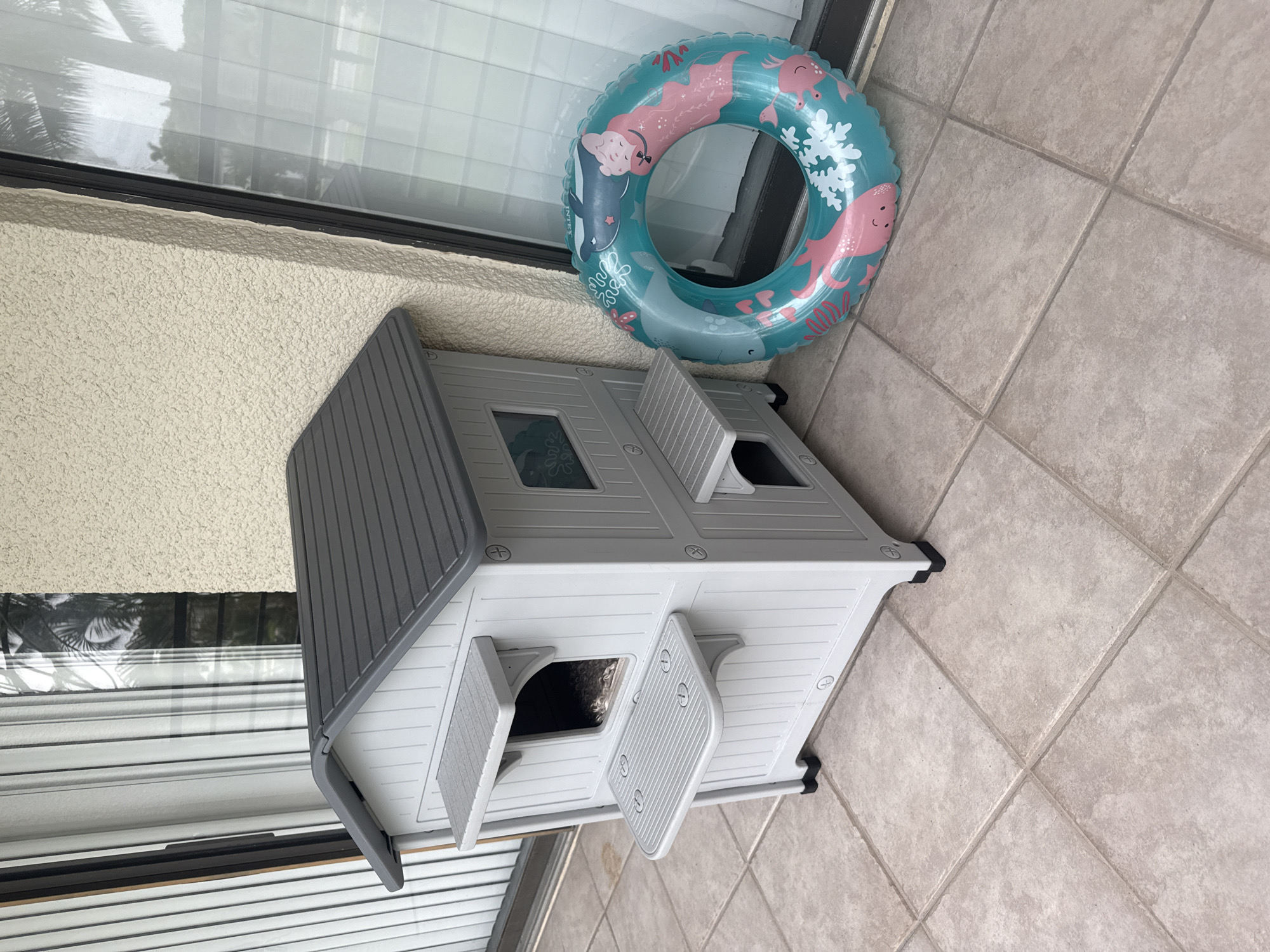 An outdoor patio scene in Boca Raton, United States features a two-story grey plastic cat house with grey sloped roof and attached platforms. A teal inflatable swim ring with a marine life print, including whales, octopuses, and coral, lies on the tiled ground to the right of the cat house. The background shows a textured beige wall and a large window with vertical blinds.