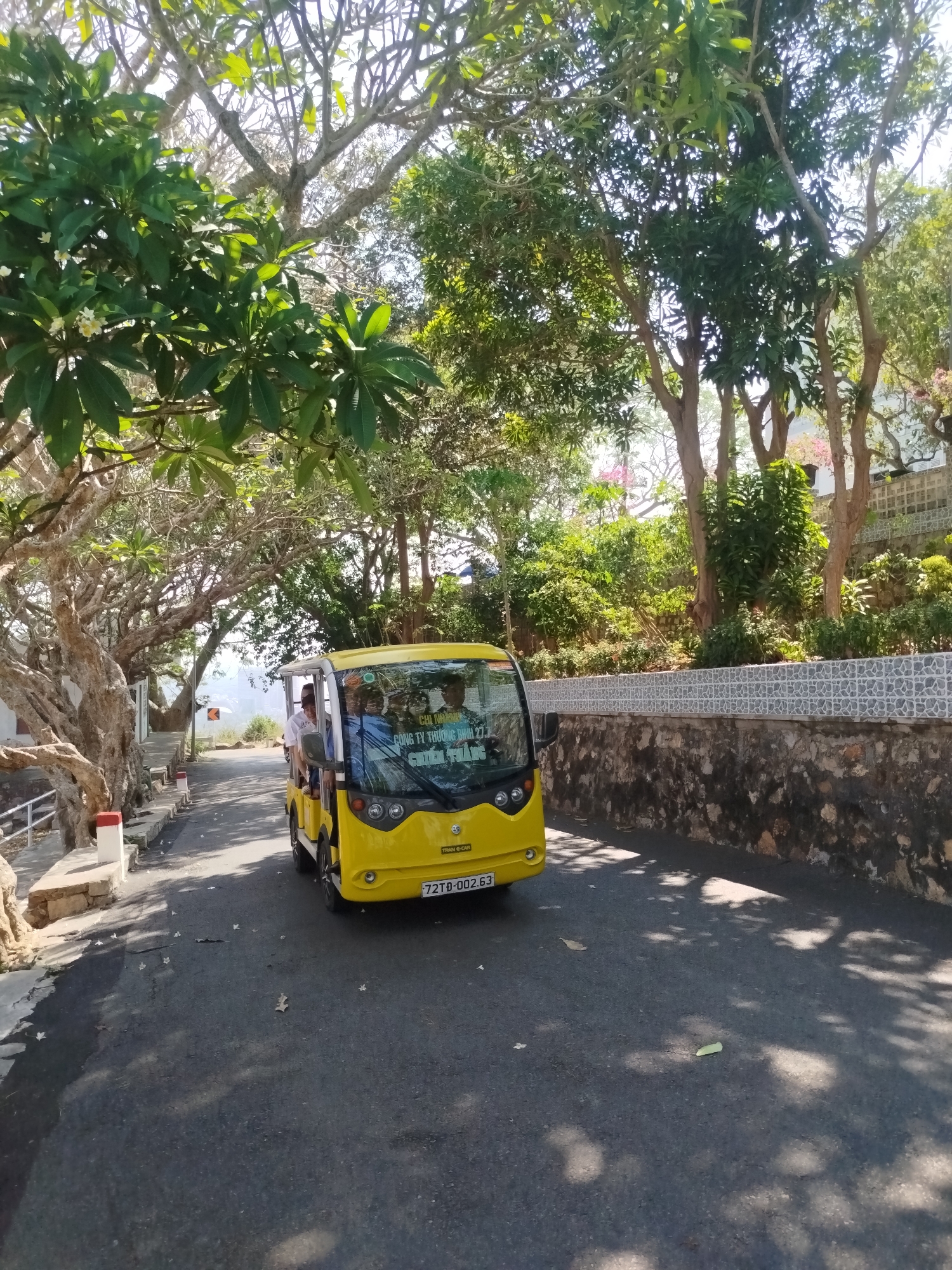 A yellow electric shuttle bus is driving on an asphalt road. People are visible sitting inside the bus. The road is lined on either side by trees and a stone wall on the right. The bus license plate reads "72TD-002.63" and text on the front of the bus includes "CÔNG TY THƯƠNG BINH 27.3" and "CHIẾN THẮNG". The surroundings appear to be a park or tourist area. The location is Ho Chi Minh City, Vietnam.