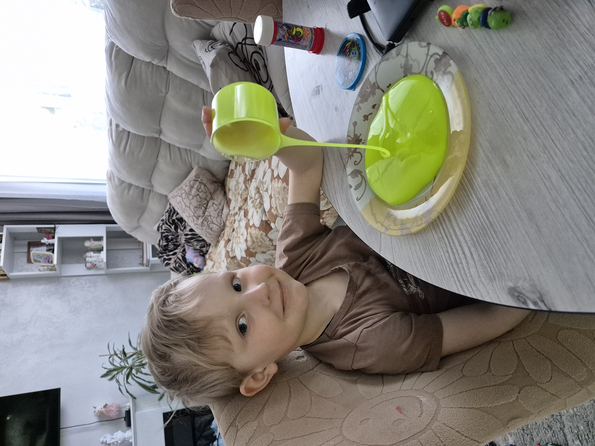 A child is sitting at a table in what appears to be an indoor setting, possibly a home. The child, with blond hair and fair skin, is wearing a brown t-shirt. The child is looking towards the camera with a slight smile. A plate containing green liquid is placed on the table in front of the child. The child is holding a green plastic cup, seemingly pouring or playing with the green liquid. Other objects on the table include a bottle of bubble solution and a row of caterpillar toys. The background shows a couch, shelves with various objects, and a plant. This image was taken in Chaikovsky, Russia.