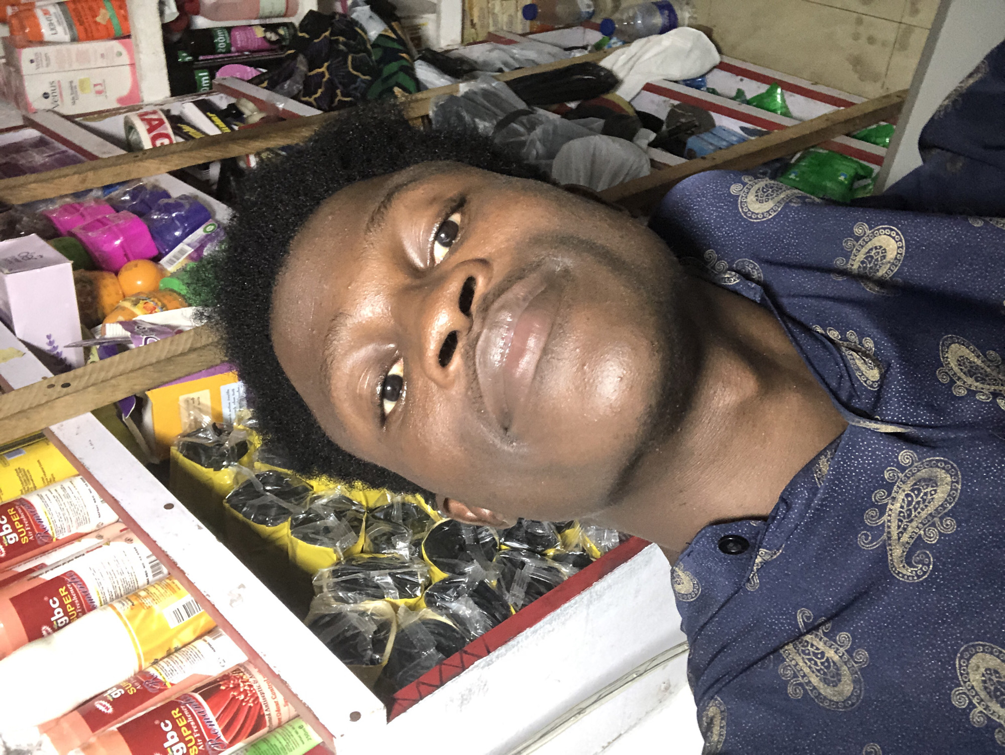 A young Black male is shown from the chest up, in a close-up, high-angle selfie. He is looking directly at the camera with a neutral expression. He has dark, curly hair and dark eyes. He is wearing a dark blue shirt with a gold paisley pattern. Behind him, there are shelves filled with various products, including aerosol cans, bottles, and plastic-wrapped items. The overall environment appears to be a retail store, possibly a small shop, with merchandise densely packed on shelves. The lighting is somewhat dim, suggesting an indoor setting. The context of Ado, Nigeria, suggests this could be a shop in a marketplace or commercial area within the city.