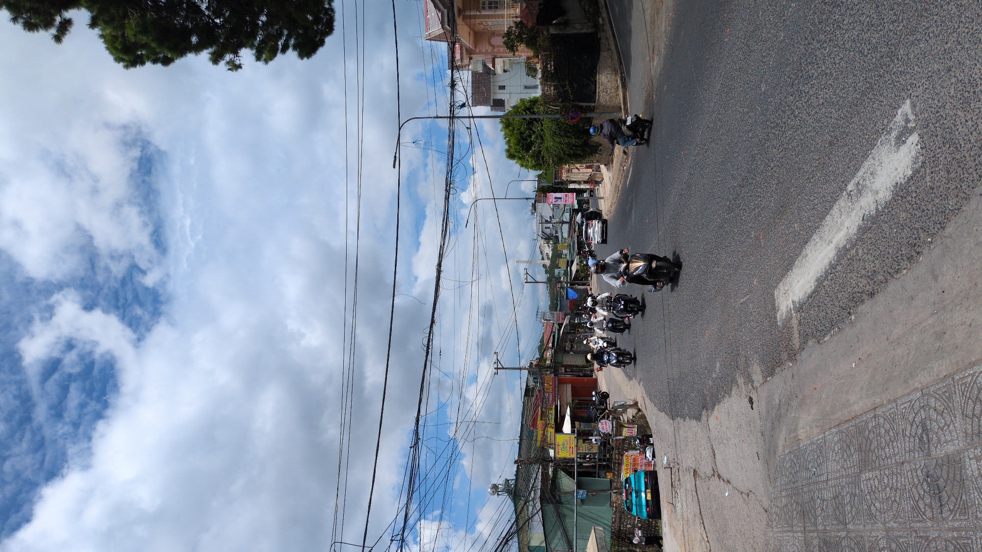 The image depicts a street scene in Da Lat, Vietnam, on a partly cloudy day. A convoy of motorcycles is traveling down a paved road. The motorcycles are of various colors and models, with riders wearing helmets and casual clothing. A few cars are also visible on the road, including a light blue car parked on the left side.
The street is lined with buildings on both sides, featuring a mix of residential and commercial structures. Some buildings have signs in Vietnamese, indicating shops or businesses. Overhead, a complex network of electrical wires crisscrosses the sky, a common sight in Vietnamese cities.
On the left side of the road, a sidewalk with decorative tiles is present, and further back, a slope leads up to a building. On the right side, there are more buildings, including a multi-story house with a pink facade. Green trees and foliage are interspersed throughout the scene, adding to the natural beauty of Da Lat. The overall atmosphere is one of a bustling urban environment with a touch of natural scenery.