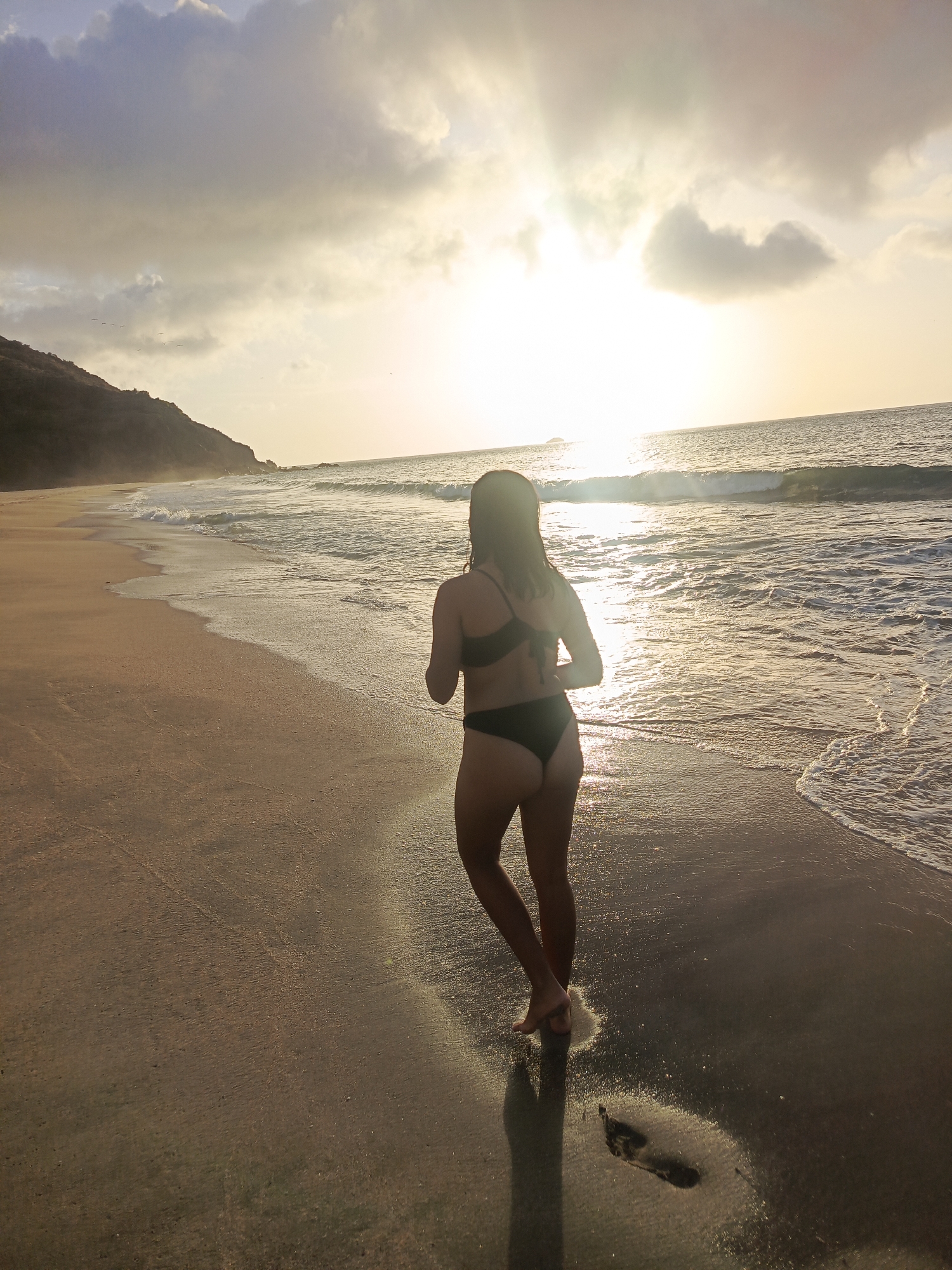 A lone individual, silhouetted against the setting sun, walks along the shoreline of Pedro González, Venezuela. The person, wearing a dark bikini, is facing away from the camera, their feet leaving imprints in the wet sand. The ocean stretches out before them, with gentle waves lapping at the shore. The sky is a mix of clouds and sunlight, creating a warm, golden glow. A rugged coastline rises in the distance, adding a dramatic backdrop to the serene scene.