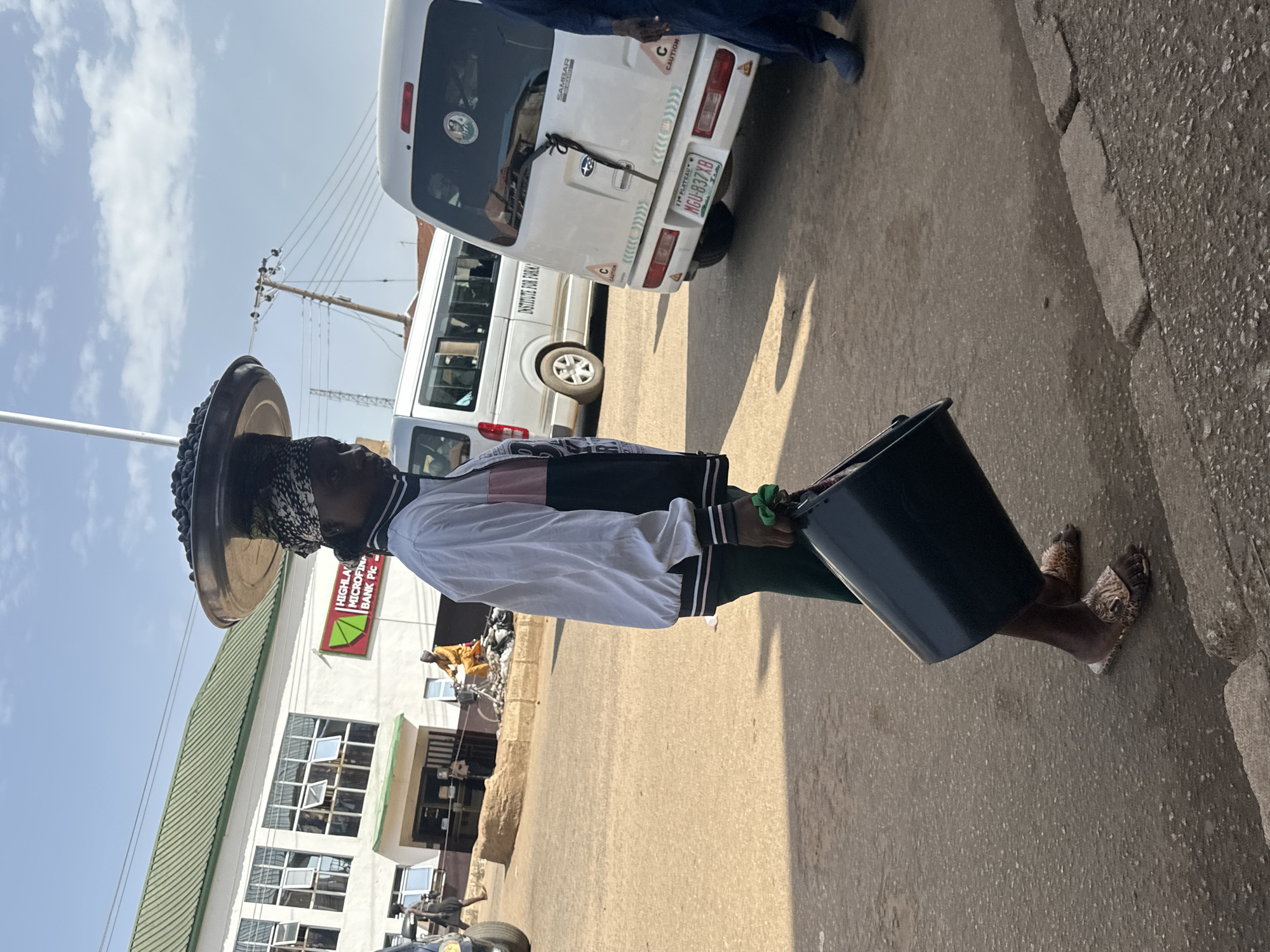 A woman is walking on a street in Bukuru, Nigeria, carrying a large black bucket in her right hand and a tray of dark berries balanced on her head. She is wearing a black and white jacket with pink accents, dark green pants, and patterned slip-on sandals. A white van with the text "INSTITUTE FOR..." visible on its side is parked behind her. In the background, a white building with a green roof has a sign that reads "HIGHLAND MICROFINANCE BANK PIC". Several other vehicles and people are visible in the distance, suggesting a commercial or urban setting. The ground is paved with asphalt, and there is a concrete curb along the right side of the street. The sky is clear and blue with some scattered clouds.