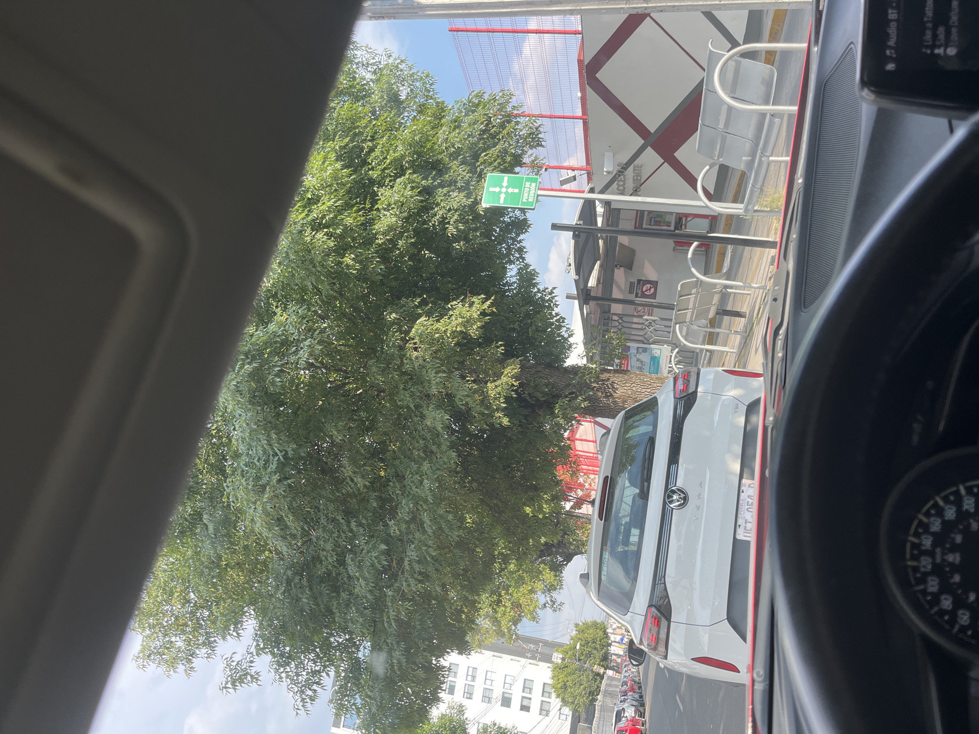 The image, taken from inside a vehicle, shows a traffic scene in Puebla, Mexico. A white Volkswagen Taos with license plate “UFT-054-B” from Puebla is directly in front of the camera car. A bus stop with waiting benches, a meeting point sign ("PUNTO DE REUNION"), and an "ACCESO PONIENTE" sign can be seen to the right. A large tree is visible to the left of the frame, and buildings are visible in the background. The vehicle's speedometer is visible on the right side. The audio system display shows "Audio BT-". The image is taken during the daytime.