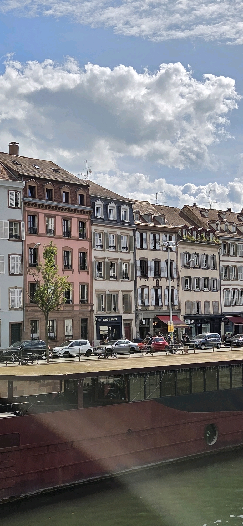 The image shows a street scene in Strasbourg, France, likely along the canals or a riverfront. In the foreground, a large, dark red boat or barge occupies the lower half of the frame, with its hull reflecting in the green water below. The boat appears to be moored or docked.
Behind the boat, a street lined with multi-story buildings stretches across the middle ground. The architecture suggests a European city, with distinct window styles, shutters, and rooflines featuring dormer windows. The buildings vary in color, with one prominently featuring a pale pink facade, another a light beige, and others lighter muted tones. Several businesses are visible on the ground floor, including a sign for "bang burgers." Cars are parked along the street, and a few individuals are depicted, some walking and one person riding a bicycle. A tree with sparse foliage stands in front of one of the buildings.
The upper portion of the image displays a bright blue sky filled with dramatic, puffy white clouds. The overall impression is a sunny day in an urban setting with historical architecture.