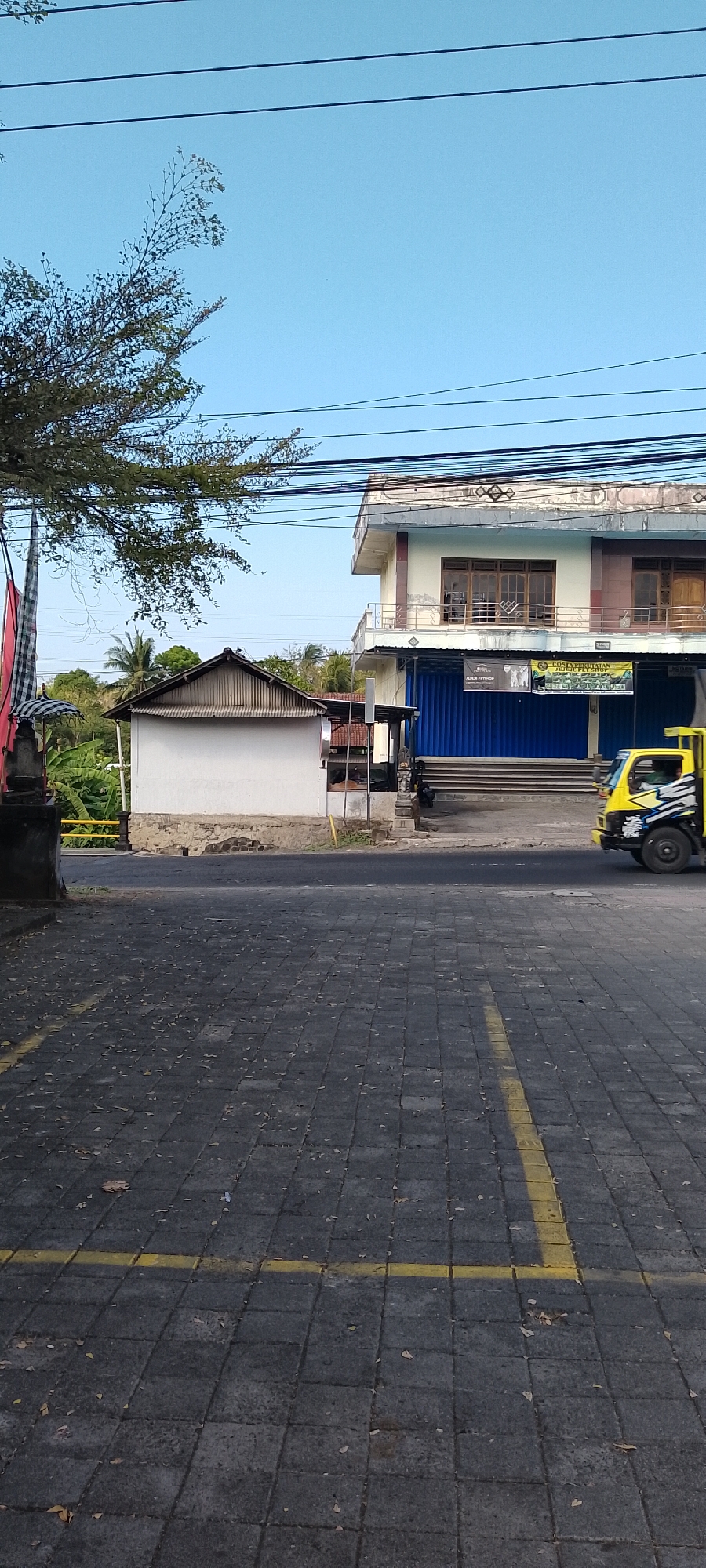 The image depicts a street scene in Pekutatan, Indonesia. In the foreground, a paved area with dark gray interlocking pavers is marked with yellow lines, suggesting a parking or pedestrian zone. Fallen leaves are scattered across the pavers. A road runs parallel to this paved area. On the right side of the road, a yellow and black truck is in motion, facing towards the right of the frame. Behind the road and pavers, there are several buildings. To the left, a small, single-story building with a corrugated metal roof and white walls stands. Next to it, a larger, multi-story building with a light-colored facade, brown-framed windows, and a balcony is visible. The lower level of this building features blue shutters. A banner with text and an image is displayed on the front of this building. Further to the left, a flagpole with a red and black striped flag and a traditional Balinese umbrella is positioned near a decorative stone element. In the background, lush green vegetation, including palm trees, is visible, and the sky is a clear, bright blue. Overhead, multiple power lines crisscross the sky.