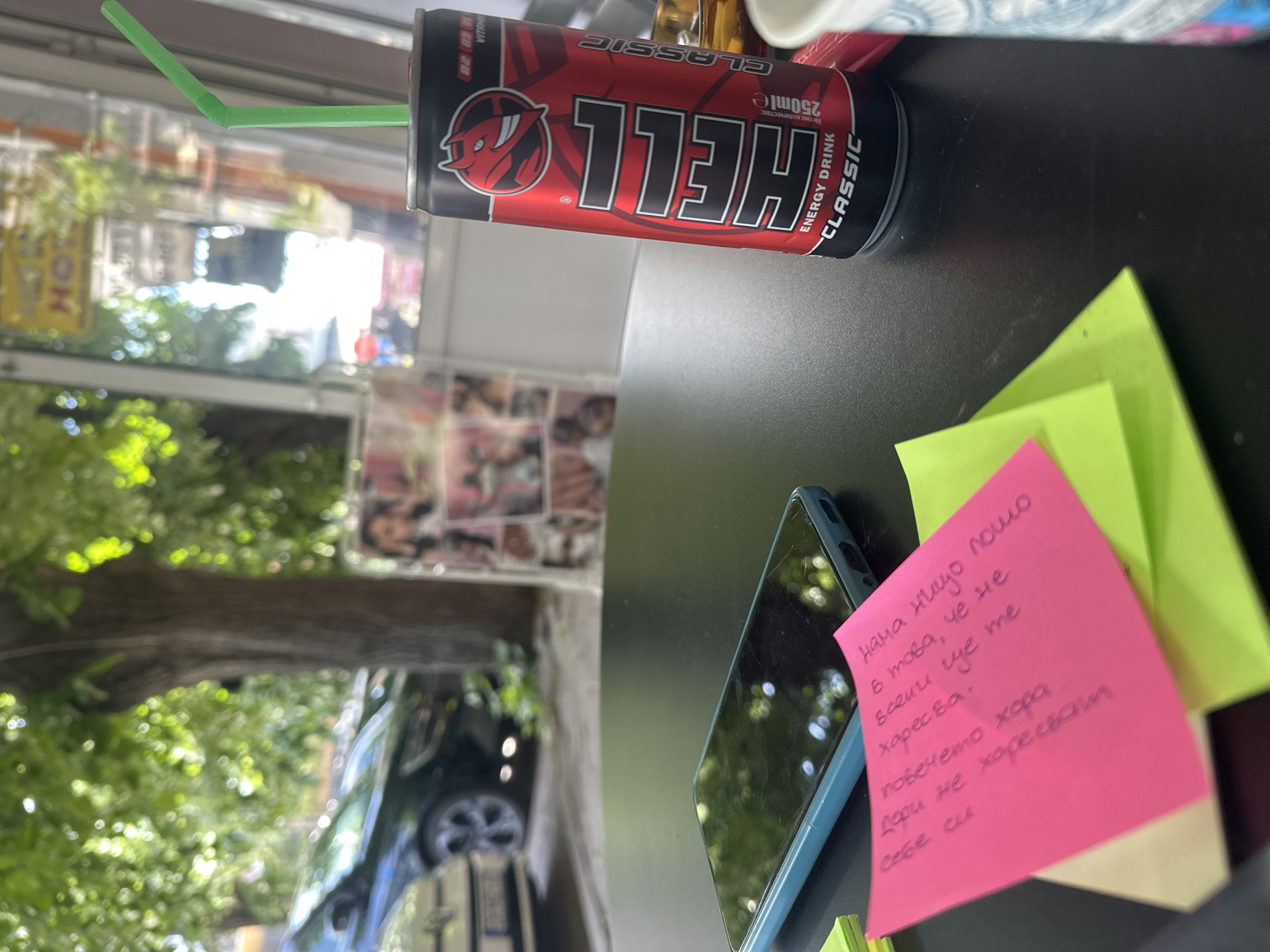 The image shows a desk or table with several items: a can of "Hell" energy drink with a green straw, a mobile phone with a cracked screen, and a stack of sticky notes, one of which is pink and contains text in Bulgarian. Outside, visible through a window, are trees, a parked car, and a building or shop front. The location is possibly in Veliko Tarnovo, Bulgaria.