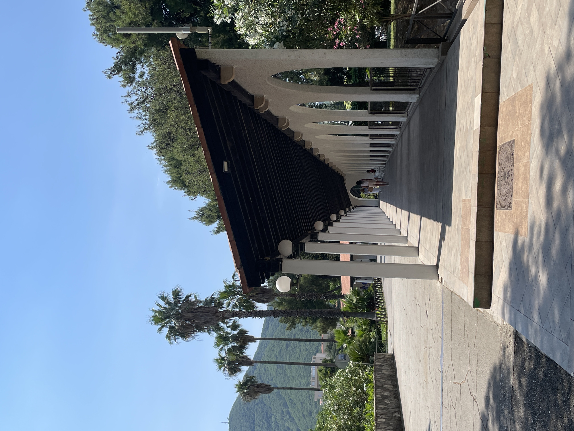 The image depicts a pedestrian walkway with a covered colonnade in Budva, Montenegro. The colonnade features a series of white pillars supporting arched openings and a dark brown, gabled roof. Spherical light fixtures are mounted along the underside of the roof.
The walkway is paved with light gray stone tiles and bordered by a low wall on one side. Two women are visible walking in the distance. In the background, palm trees, lush greenery, and a mountain are seen under a clear blue sky.