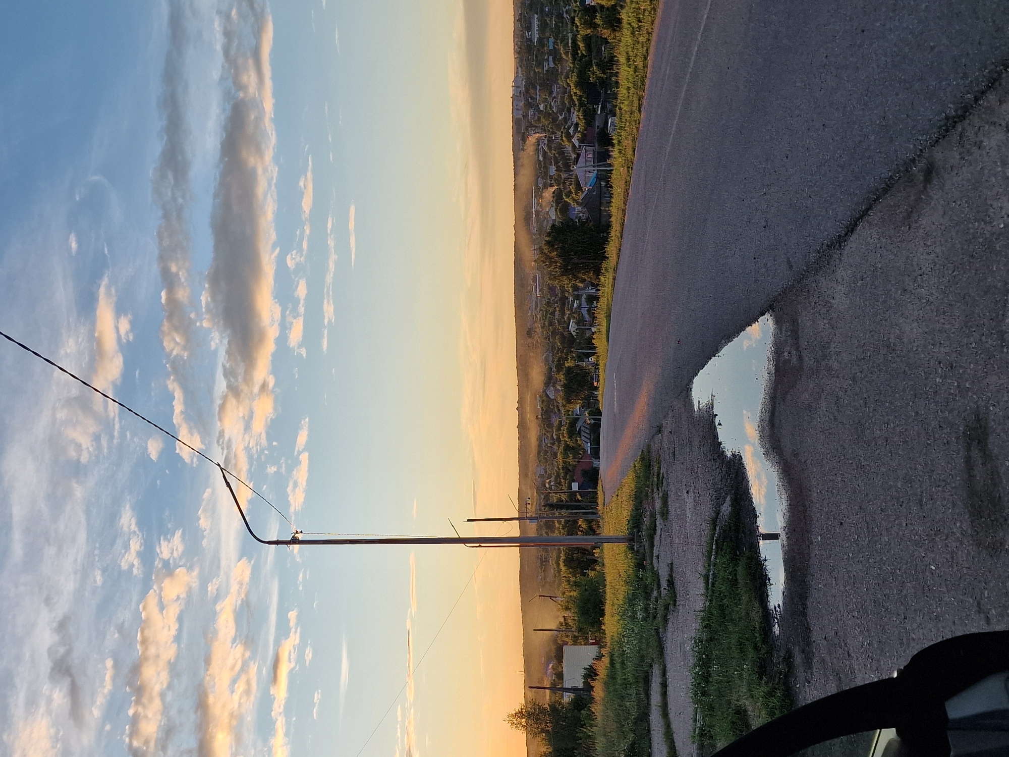 This is a vertical, high-angle, outdoor, eye-level shot of a rural landscape at sunset. The scene is divided horizontally between the sky and the ground. The sky, occupying the left half of the frame, is filled with scattered, wispy clouds tinged with orange and yellow from the setting sun. The clouds are set against a pale blue sky. A thin black wire runs diagonally across the sky.
The right half of the frame depicts a rural village and a paved road. The village is nestled in a valley, with numerous houses and buildings visible amidst dense greenery and trees. The road, in the foreground, is dark asphalt and appears wet, reflecting the sky and surrounding scenery. A puddle on the road further mirrors the sky. The overall impression is one of a serene, end-of-day atmosphere in a rural setting. The time of day is clearly sunset, indicated by the warm hues in the sky and reflections. There are no people visible in the image.