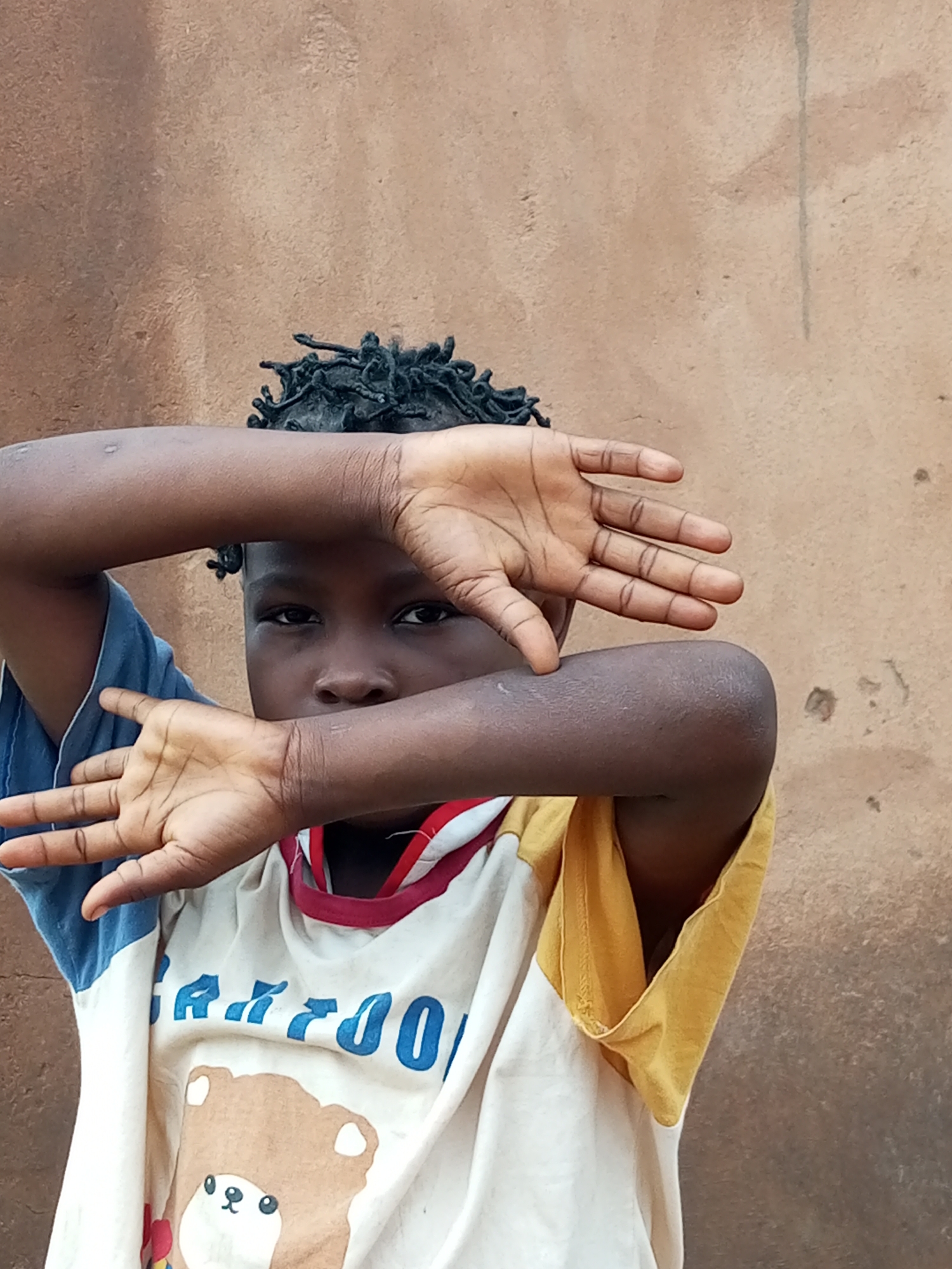 A close-up, medium shot captures a child's face peeking through their arms, which are held up in front of their face. The child has dark skin and dark, braided hair. They are wearing a t-shirt with a cartoon bear design and blue and yellow accents. The background is a plain, textured brown wall. The child's hands are positioned as if blocking the view of their face.