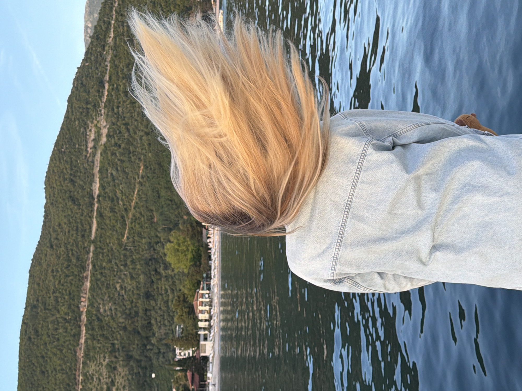 A medium shot from behind shows a person with long, light blonde hair wearing a light-wash denim jacket. The person is outdoors with a body of water in the foreground and a densely forested mountain in the background. In the distance, a row of buildings lines the shore of the body of water. The scene is in the city of Budva, Montenegro, based on the landscape and architecture visible in the image. The water has a slight ripple, and the hair is slightly disheveled, indicating a gentle breeze. A small, brown object, possibly a plush toy, is visible at the top right of the image on the person's shoulder.