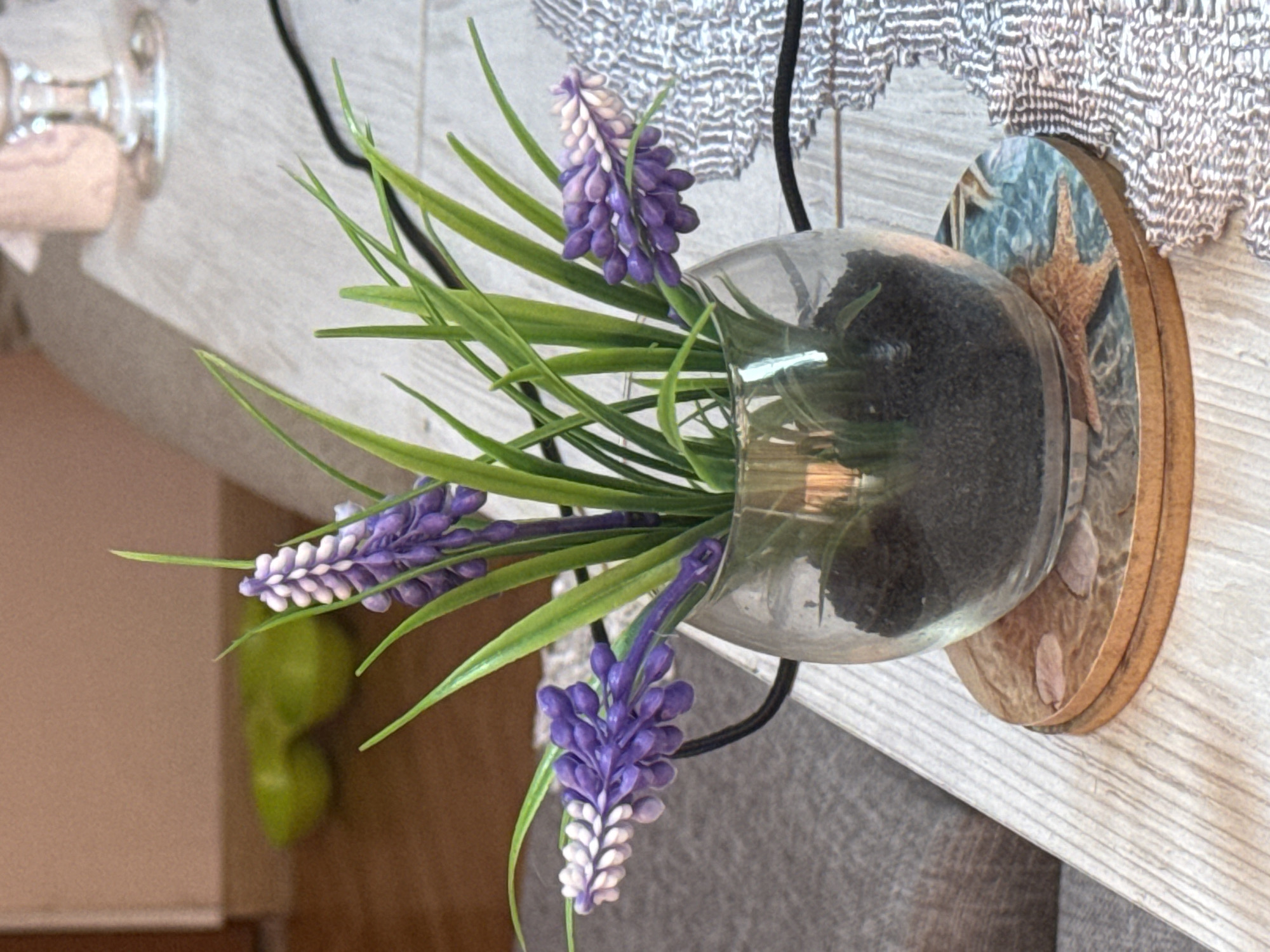 A close-up, slightly angled shot shows a small glass vase filled with dark soil and artificial purple flowers with green stems. The vase is placed on a circular coaster depicting a marine scene with a starfish. The vase and coaster sit on a light-colored wooden surface. In the background, a portion of a grey sofa and a blurred green object are visible. To the right of the vase, a patterned textile is partially visible. The scene appears to be indoors, on a table, likely in a residential setting in Montana, Bulgaria. No people are present in the image.