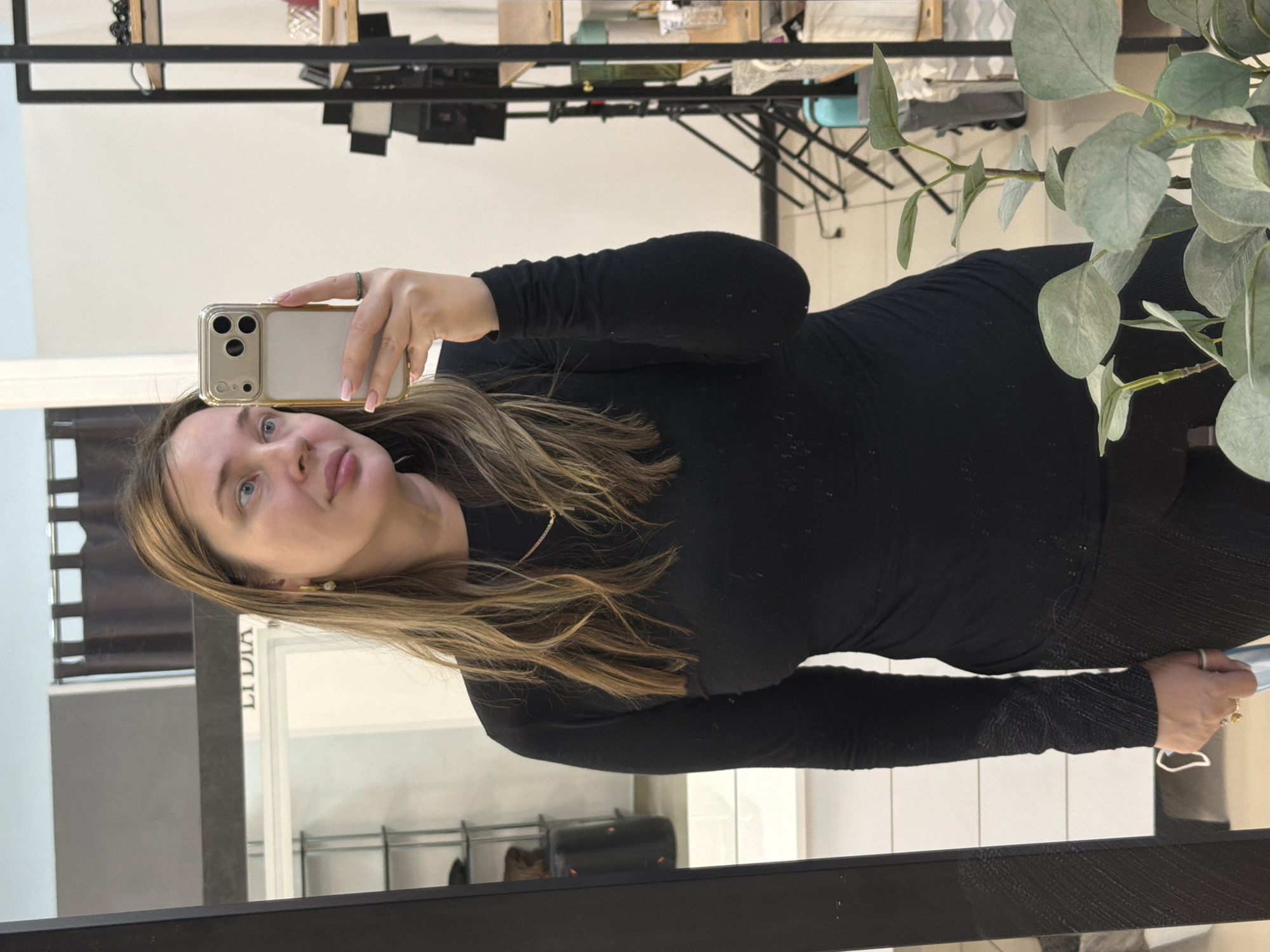 An adult female is depicted taking a mirror selfie indoors at an establishment identified as "LIDIA" in Chaikovsky, Russia. The female, positioned centrally, looks towards her reflection while holding a light-colored smartphone, equipped with a transparent gold-edged case and a triple rear camera array, in her right hand. She has long, light brown hair with blonde highlights, blue eyes, and is wearing a black long-sleeved top. Accessories include a gold chain necklace, a gold stud earring in her visible left ear, a silver ring on her left ring finger, and a green ring on her right ring finger. The reflected background, visible through a large, dark-framed mirror, shows a commercial interior with white walls, a light-colored tiled floor, a black metal shelving unit with wooden shelves holding various items, and a black professional photography light stand with attached lighting equipment. A white display unit or counter, holding a black cylindrical object, is reflected on the left, beneath a dark brown curtain. A plant with light green, eucalyptus-like leaves occupies the unreflected foreground, partially obscuring the lower right.